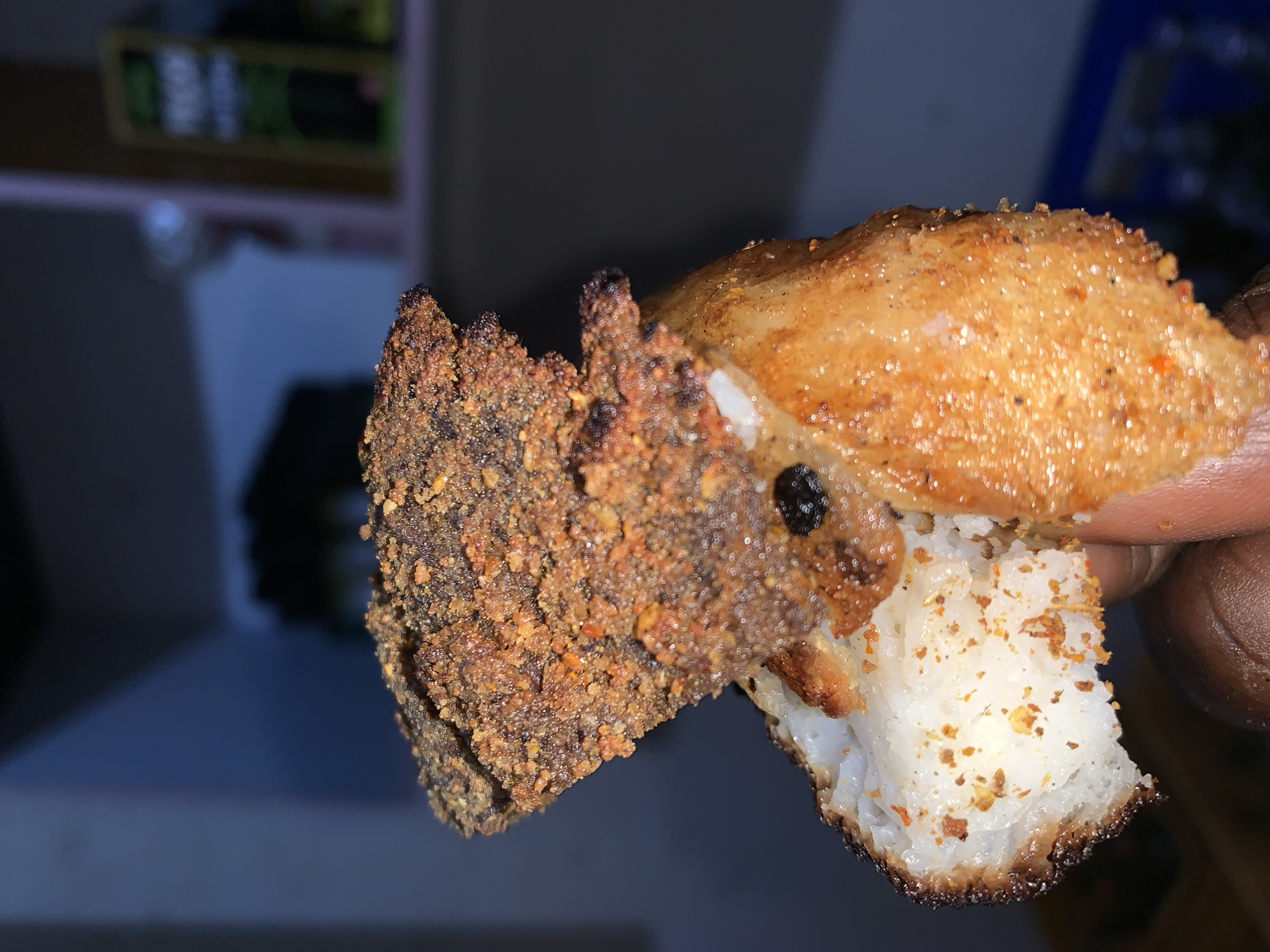 A close-up image shows a piece of food being held. The food consists of a base of white, rice-like substance, topped with a layer of seasoned, browned meat. The exterior of the meat has a crusty, textured appearance. The background is blurred, but includes indistinct shapes and colors suggesting an indoor environment. The food is being held by fingers with darker skin tone. The location in the city of New Stadium, Nigeria is not determinable from the image.