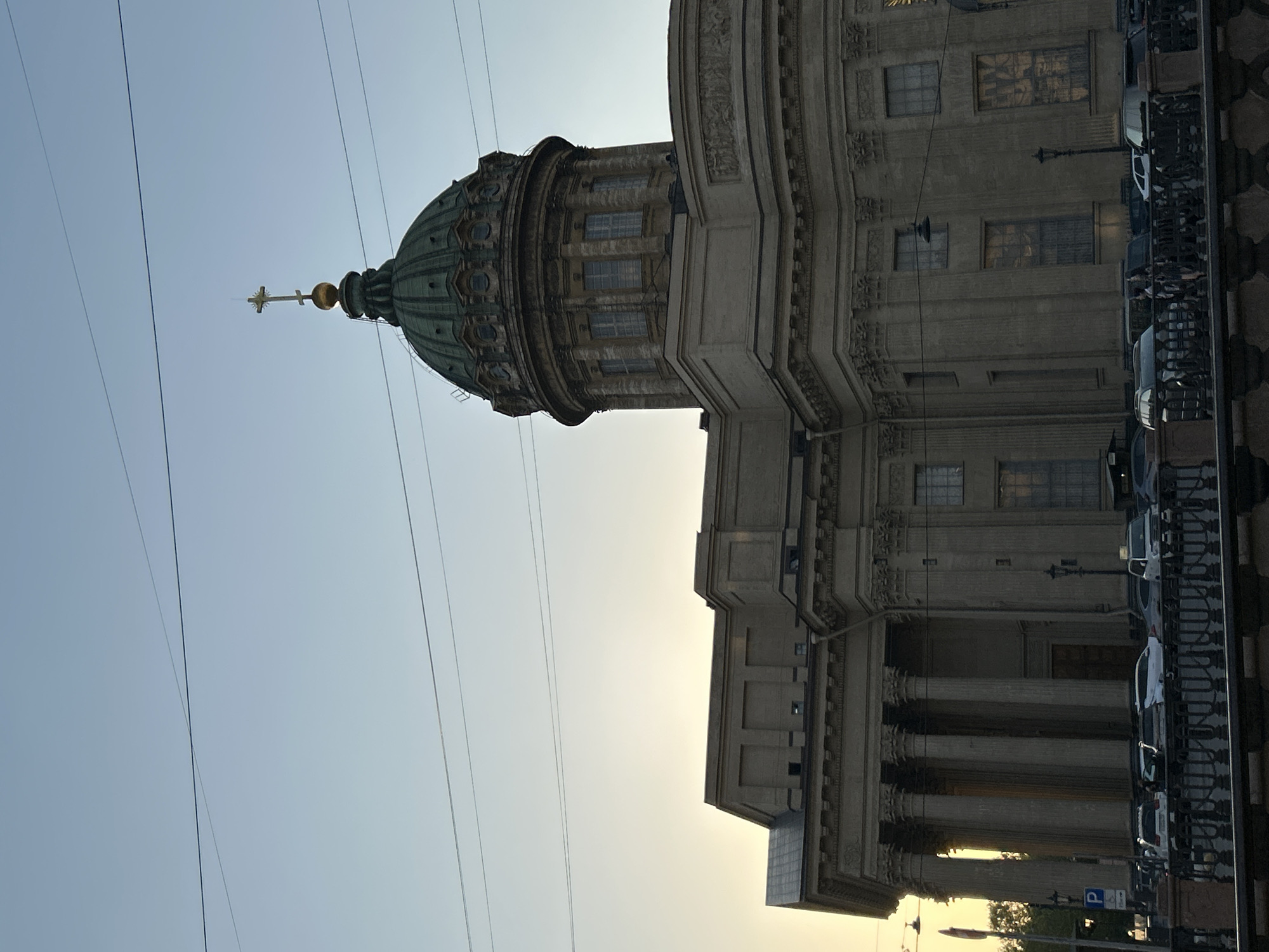 The image features the Kazan Cathedral in Saint Petersburg, Russia. The view is a low-angle shot, emphasizing the cathedral's dome, topped with a golden spire and cross. The facade of the building is visible on the right, revealing architectural details such as columns, windows, and ornate carvings. Alongside the building, parked cars line the street, adjacent to a decorative fence. Overhead wires traverse the open sky, which transitions from blue to pale yellow in the background, indicating either sunrise or sunset. The overall scene captures a moment in the urban environment of Saint Petersburg.