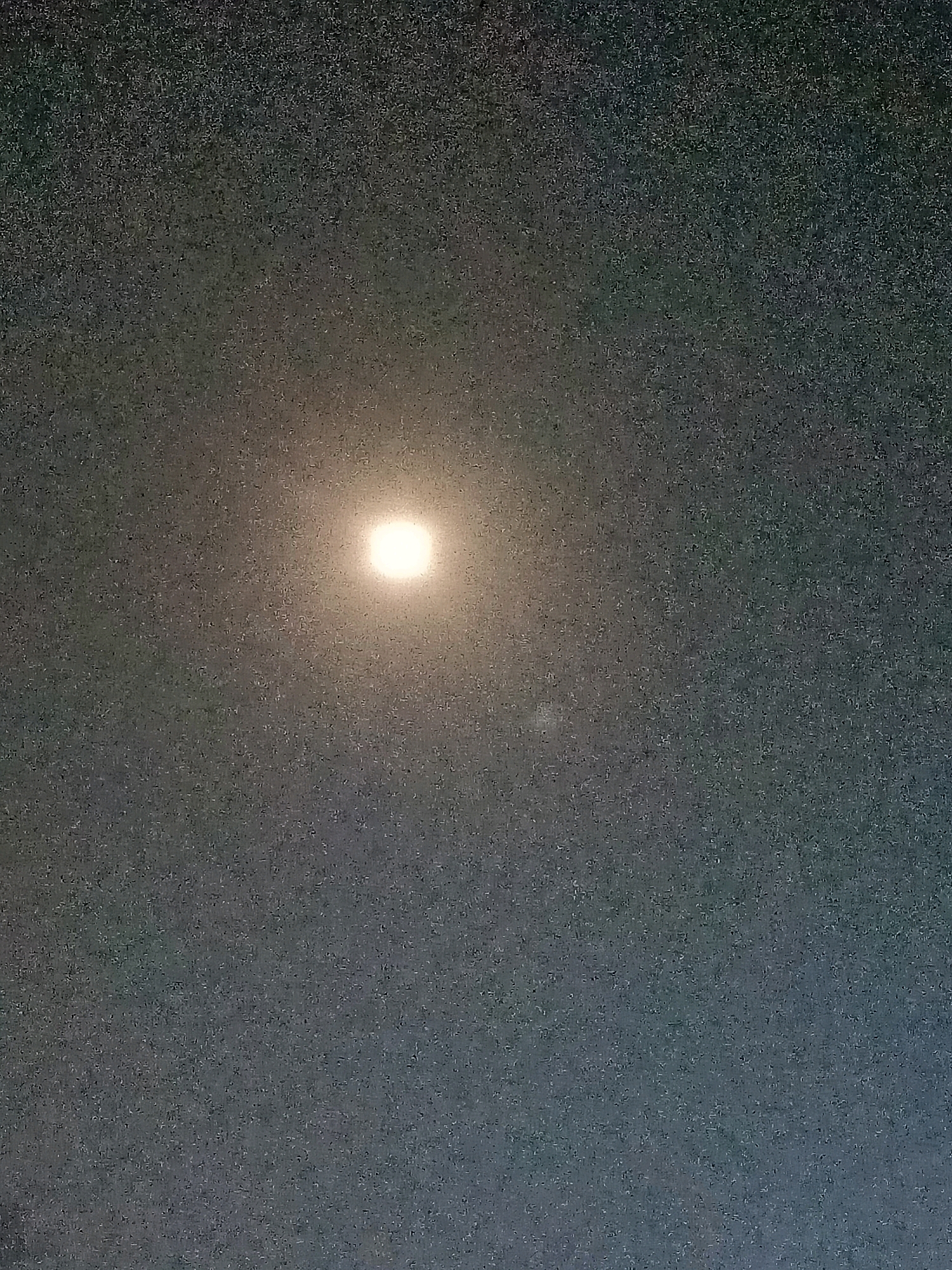 The image shows a full moon in a dark, grainy sky. The moon is a bright, circular object with a halo of light around it. The sky is a deep gray, with a faint gradient from darker at the top to lighter at the bottom. There are no other celestial bodies or objects visible in the sky. The image is taken from the perspective of someone looking upwards. The location is Badas, Indonesia.