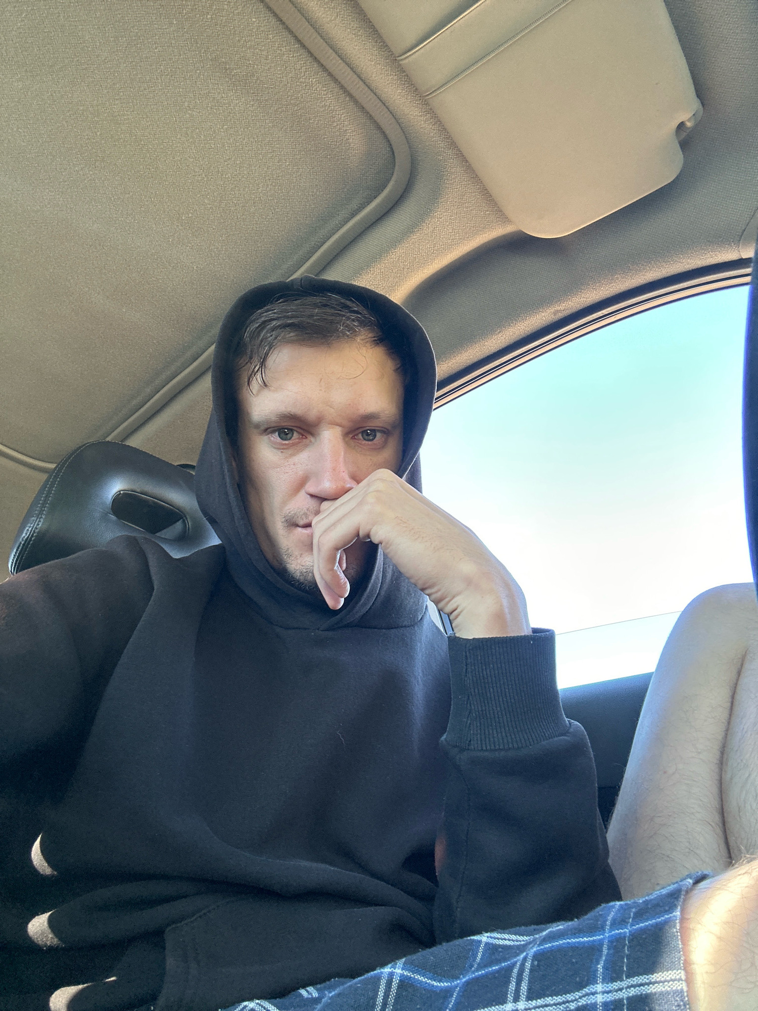 A man wearing a black hoodie and plaid pajama bottoms sits in the passenger seat of a car. He has his right hand on his chin, and his left hand is resting on his leg. The car's interior, including the headliner and a sun visor, is visible. The sky is clear and blue outside the car window. The location is not specified as being in the city of Makiv, Ukraine.