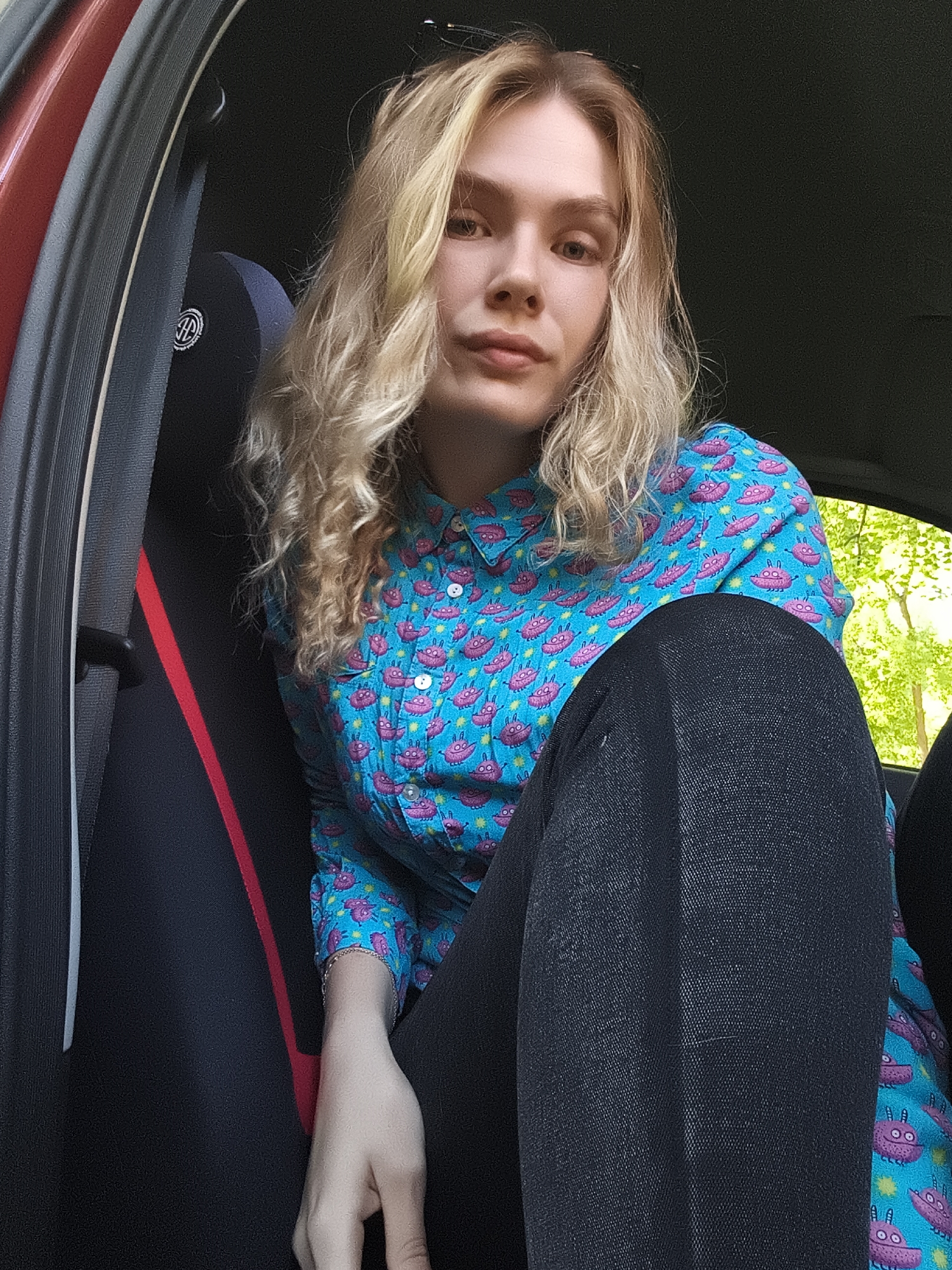 A person with blonde hair and a blue shirt patterned with pink cartoon aliens is sitting in the driver's seat of a car. The person is wearing black pants and a bracelet. The car door is open and the car seat has a red stripe. The location cannot be determined to be Izhevsk, Russia from the image.