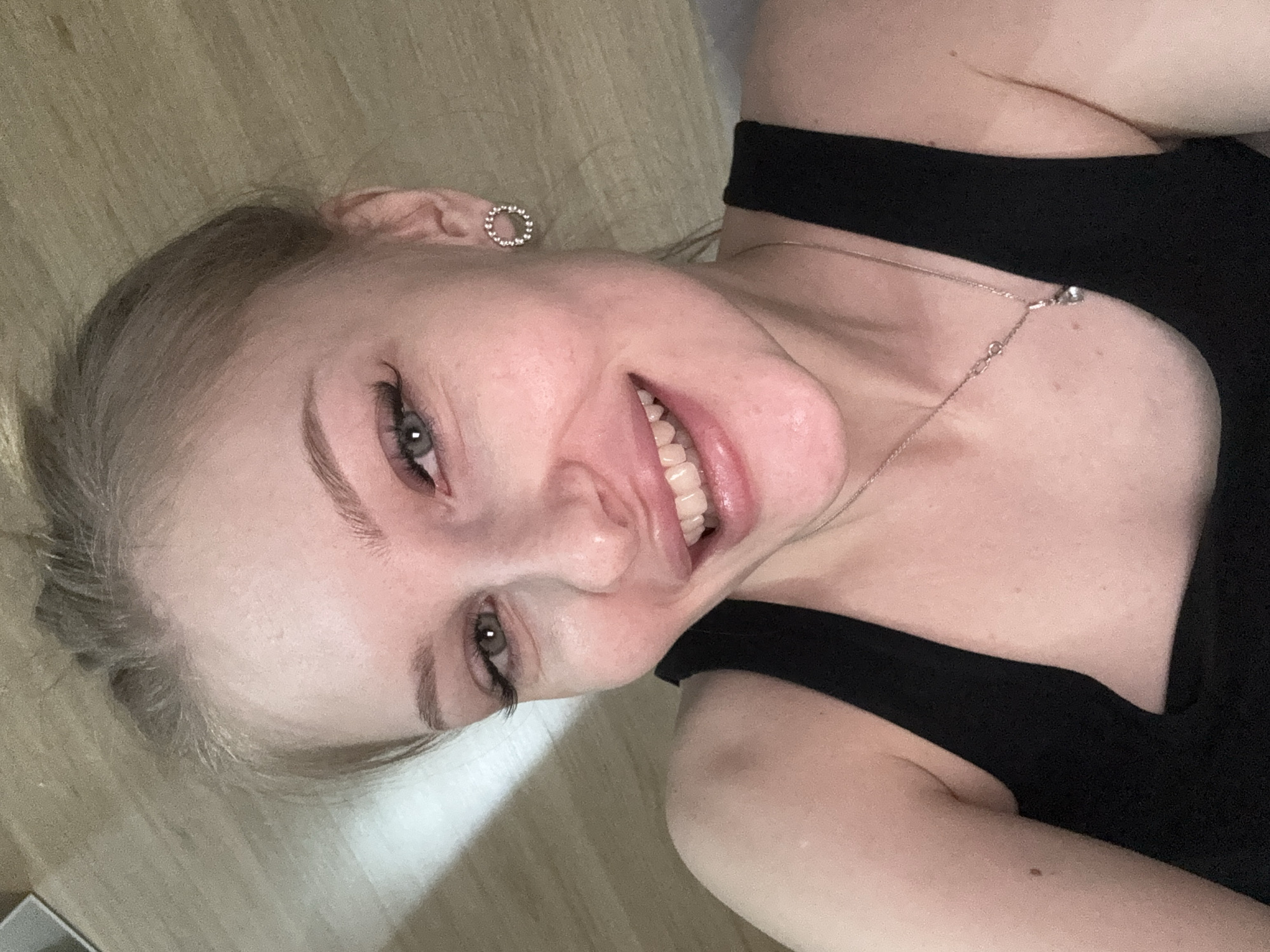A close-up shot features a young woman with fair skin, light blonde hair, and light-colored eyes. She is smiling, revealing her teeth. She is wearing a black sleeveless top. A delicate silver necklace is visible on her chest. An earring is visible on her left ear. The background appears to be a light-colored wooden surface. The scene is set indoors. There is no evidence to suggest the location in Perm, Russia.