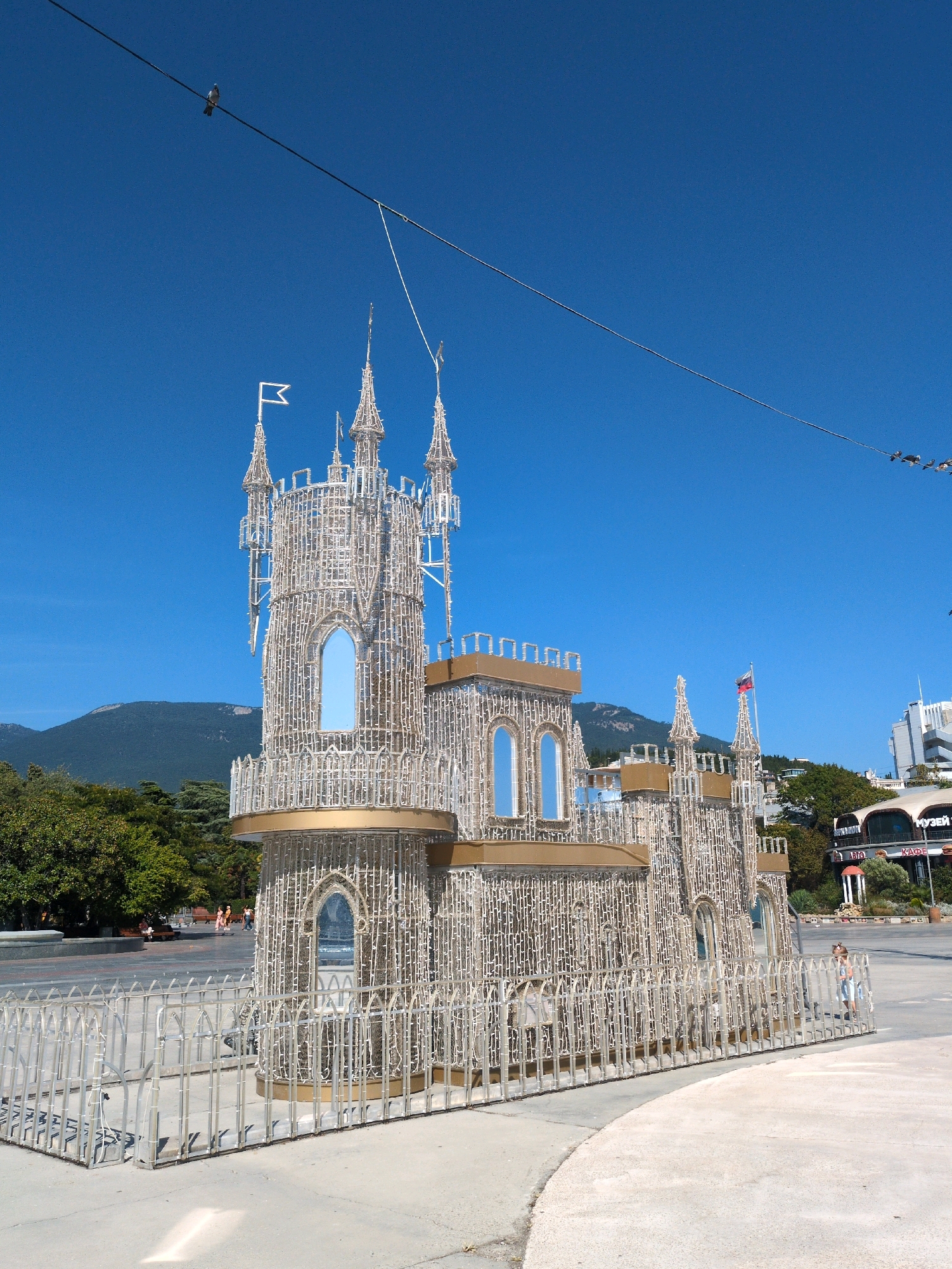 A daytime, eye-level outdoor shot shows a decorative castle structure made of lights, viewed from the side and slightly from the front. The castle has multiple towers, flags, and arched windows. It is surrounded by a metal fence. Behind the castle, green trees and mountains are visible, along with buildings and a clear blue sky. A single pigeon is perched on a wire stretching across the top of the frame. In the lower right quadrant, a young girl wearing a light-colored dress stands near the fence.