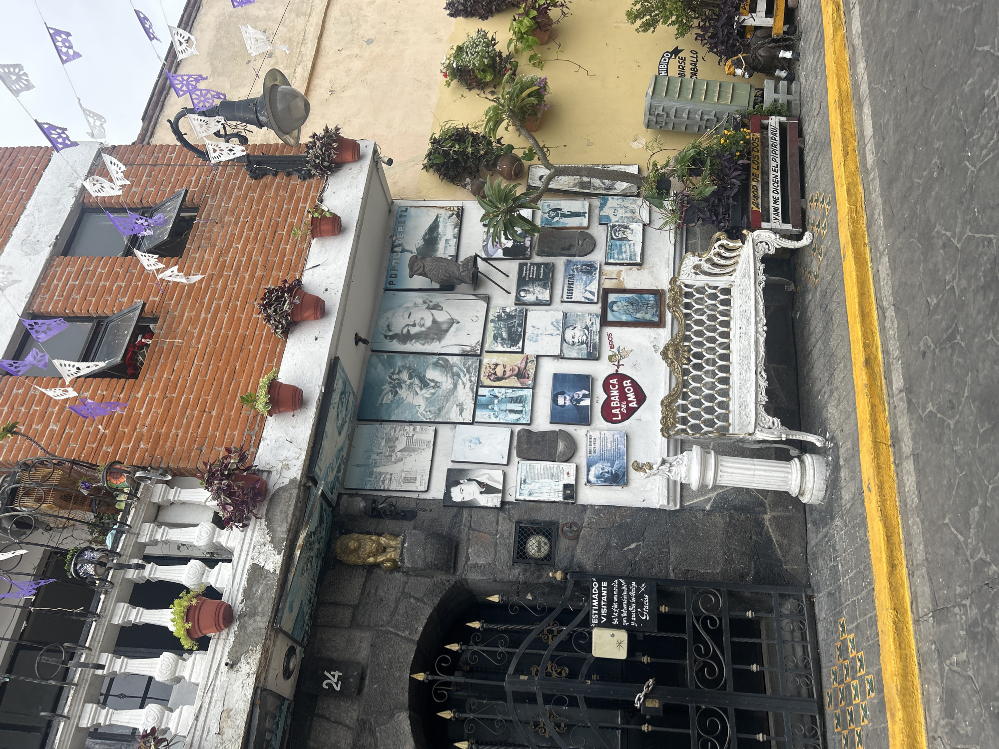 The image captures a street scene in Atlixco, Mexico, showcasing the facade of a building adorned with various decorative elements. The building's brick exterior is decorated with purple and white papel picado banners, typical of Mexican celebrations. A balcony with white balusters overlooks the street, featuring potted plants. The ground floor displays a collection of framed pictures and artwork mounted on the wall, including portraits of celebrities like Marilyn Monroe and Frida Kahlo, as well as other historical figures and scenic views. A black wrought-iron gate, decorated with ornate scrollwork, guards an entrance. Adjacent to the gate, a sign with Spanish text provides visitor information. A white, ornate bench sits on the sidewalk in front of the building, accompanied by a white pedestal. The street is paved with cobblestones and bordered by a bright yellow curb. In the background, a yellow-painted building and more greenery are visible, suggesting a vibrant urban setting. The overall impression is of a culturally rich and visually eclectic streetscape.