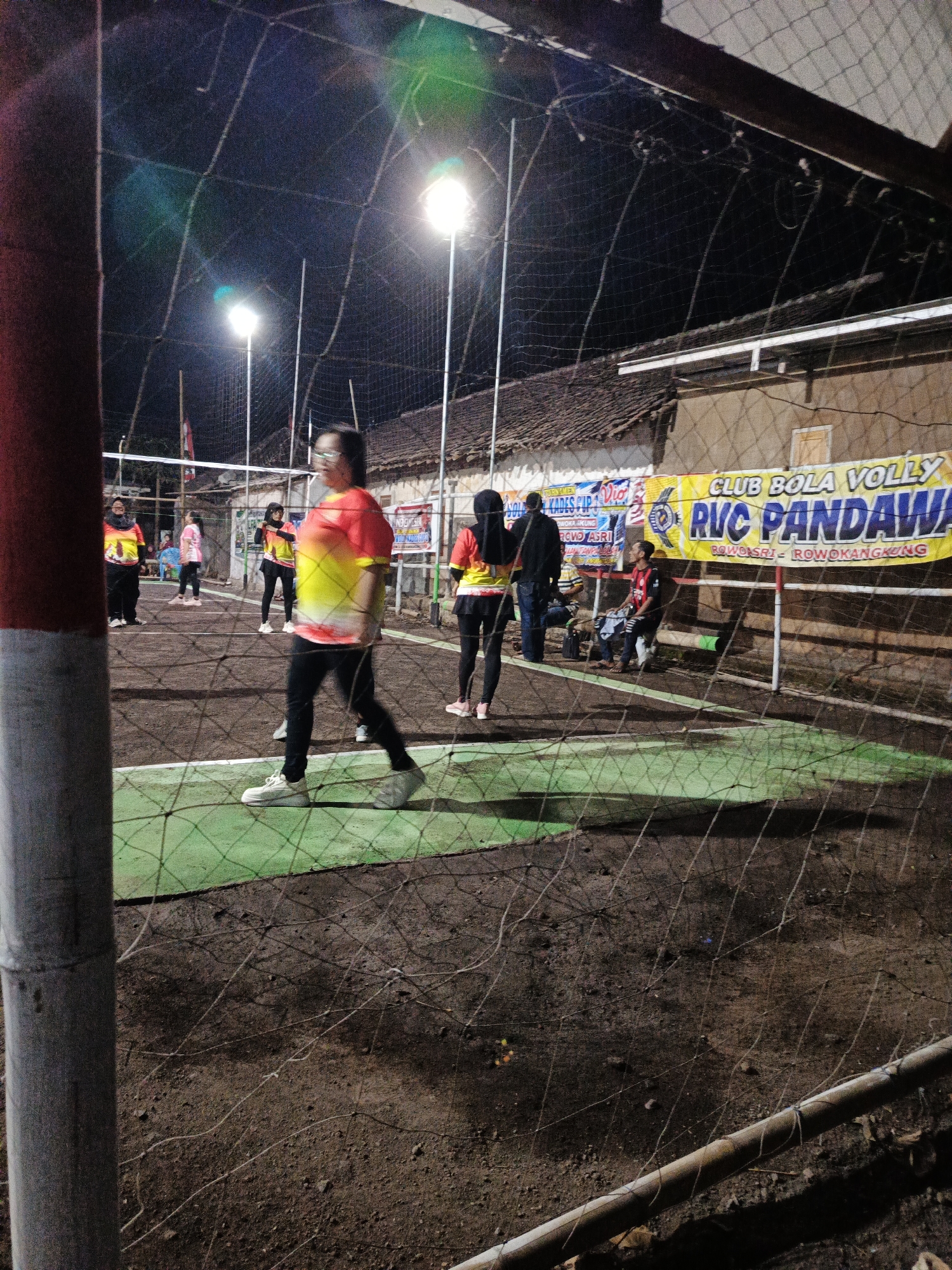 In Rowokangkung, Indonesia, an outdoor volleyball court at night is illuminated by bright lights. Several individuals are present on and around the court. A person in the foreground, facing left, wears a blurred yellow and red shirt and black pants, with white sneakers. To the left, another person in a similar yellow and red shirt and black pants stands with their back to the viewer. In the background, more people are visible. Two individuals stand near the net, one in a pink shirt and black pants, the other in a yellow and red shirt and black pants. A person in a red shirt and black shorts is seated on a bench to the right. A banner hangs above the court, reading "CLUB BOLA VOLLY RVC PANDAWA ROWOBASRI - ROWOKANGKUNG." The court surface appears to be a mix of green material and dirt. A net stretches across the court, and a wire fence encloses the area.