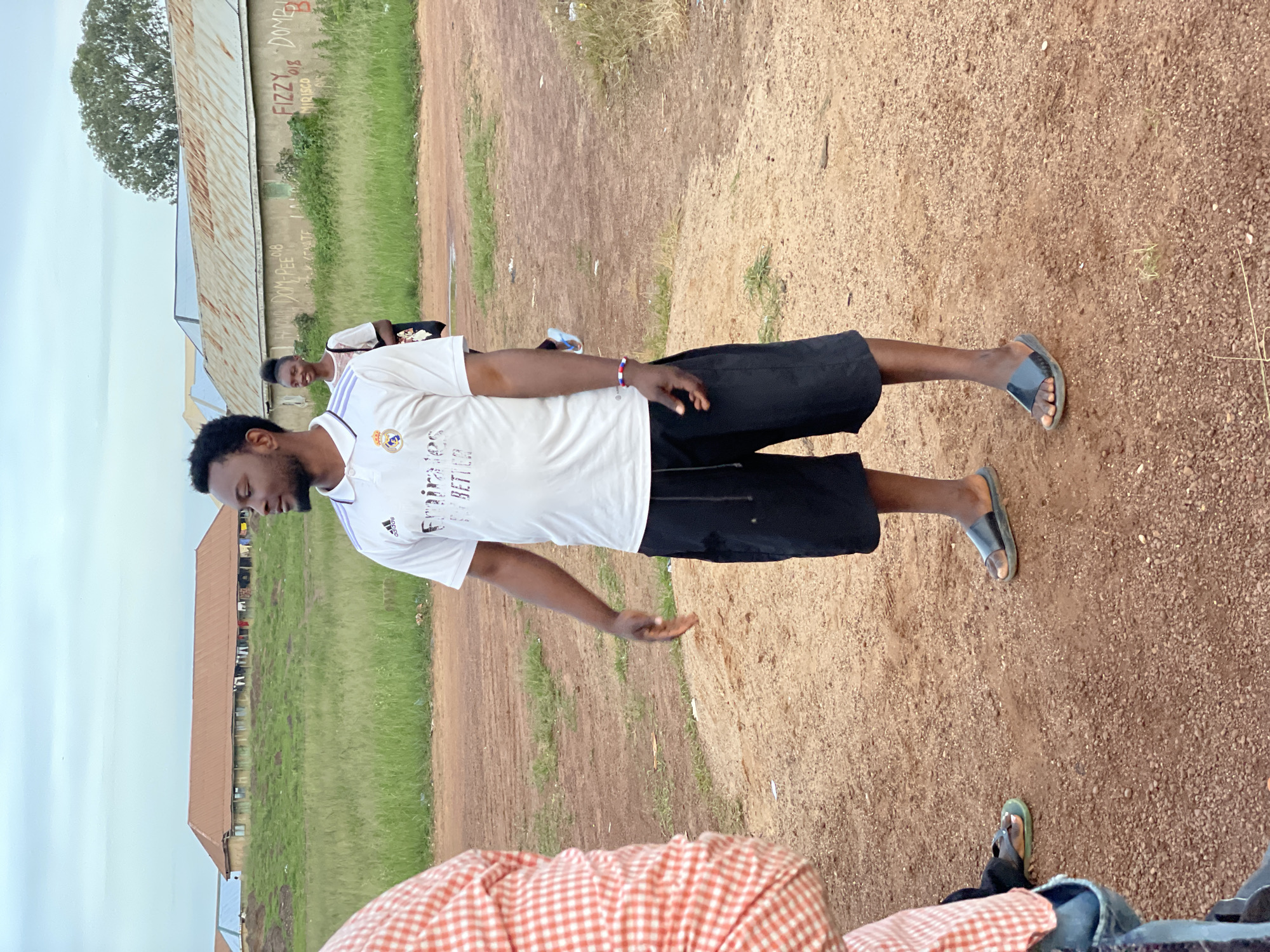 In the city of Durumi, Nigeria, an image captures a man standing on a dirt path in an outdoor setting. He is wearing a white polo shirt with the Adidas and "Emirates Fly Better" logos, and black shorts. A red, white, and blue bracelet adorns his left wrist. To his left, another person, only partially visible, wears a red and white checkered shirt. In the background, a young woman with her hair in a bun is walking, and behind her, buildings with corrugated metal roofs are visible, some adorned with graffiti. The ground is a mix of reddish-brown dirt and scattered gravel. Green grass and some scattered debris are also present in the environment. The sky above is overcast with a pale hue.