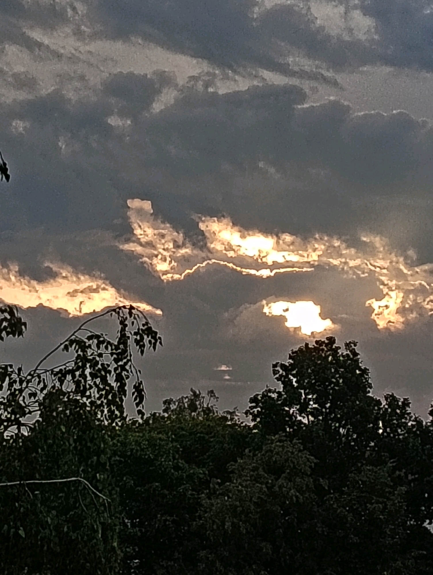 The image shows a dramatic sky with dark, heavy clouds interspersed with bright shafts of sunlight. The sunlight pierces through the clouds, creating an effect of glowing edges and illuminated patches, suggesting either sunrise or sunset. In the foreground, the tops of dark green trees are visible, their foliage silhouetted against the sky. The scene appears to be an outdoor environment, possibly a park or a residential area on the outskirts of the city of Hrodna, Belarus, characterized by the natural elements of trees and the sky. There are no people or specific urban structures clearly identifiable in the frame, making the focus solely on the natural landscape and atmospheric conditions.