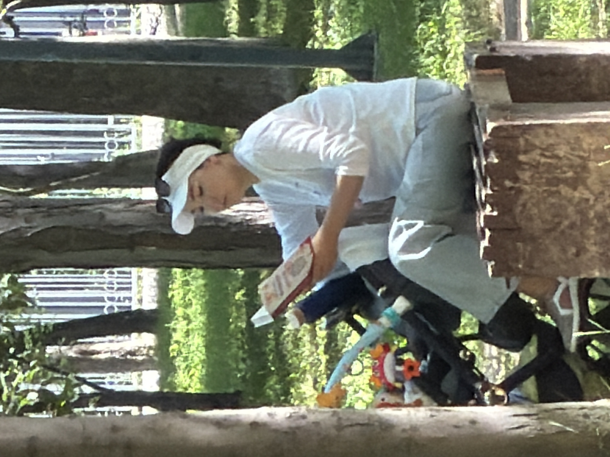 A young woman wearing a white visor and white long-sleeved shirt is reading a book. She is sitting on a wooden structure in a park. A stroller with stuffed animals is next to her. The background shows trees and a white fence.