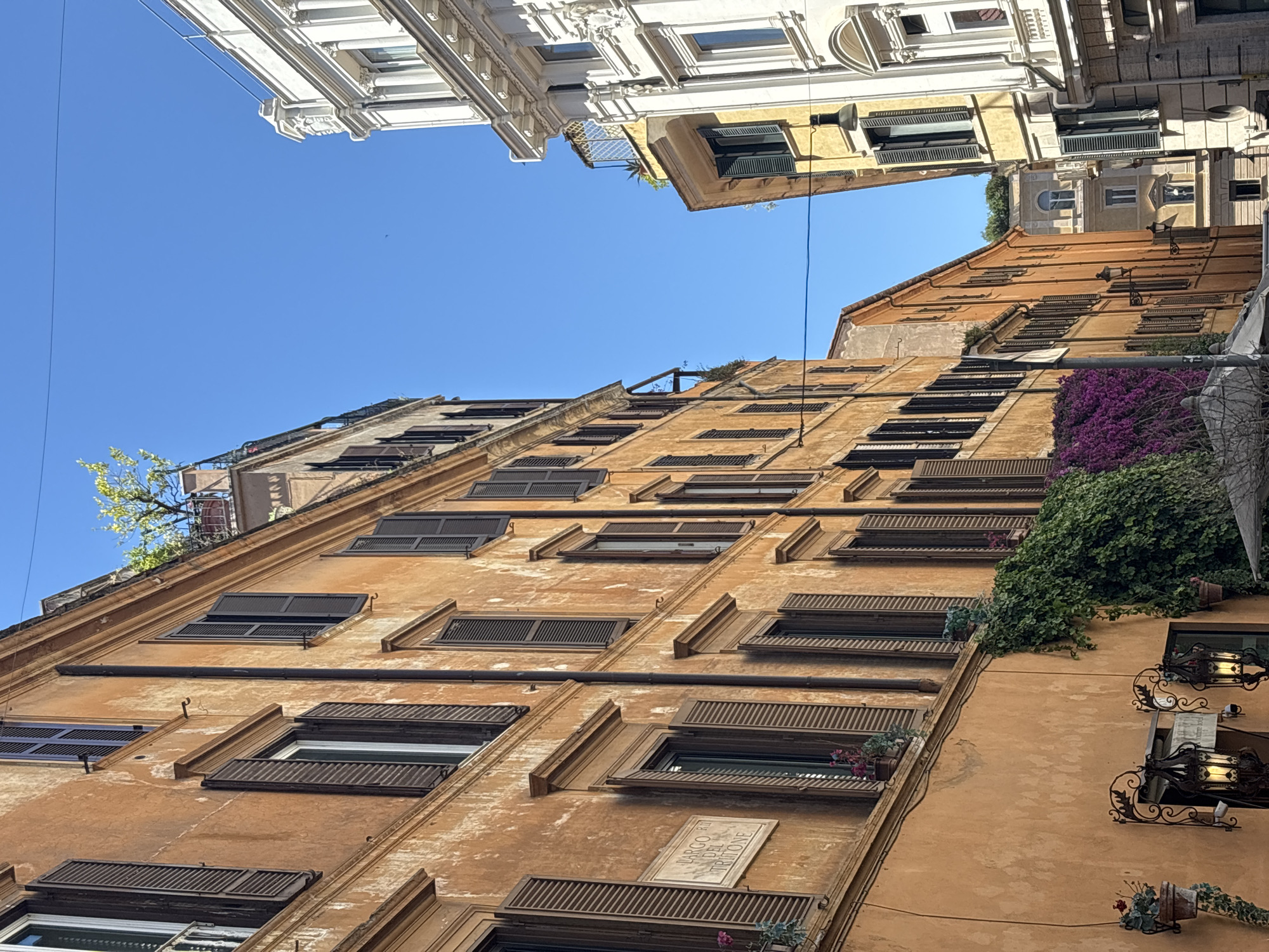 A low-angle shot captures the facade of a building in Rome, Italy, under a clear blue sky. The building has numerous windows with dark shutters and elaborate frames. Some windows feature flowerpots with colorful blooms. A plaque on the building is visible, possibly indicating a historical or cultural significance. Ivy and other plants grow on the building's exterior. Above, the upper stories of adjacent buildings are partially visible. Architectural details such as cornices and window styles suggest the area's age and historical character.