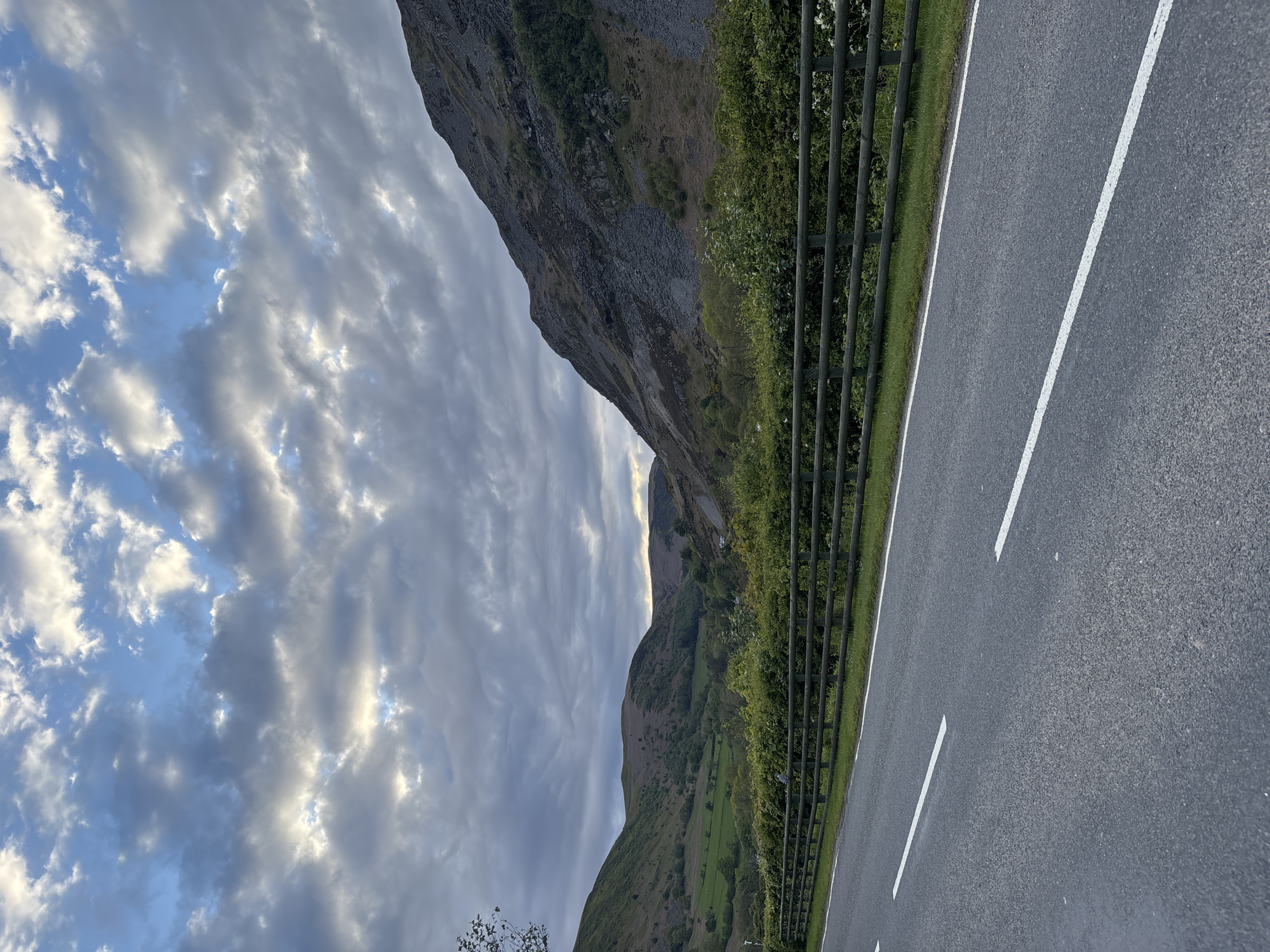 The image shows a road in Llangynog, United Kingdom, bordered by a metal safety fence and grass on one side and a mountain on the other. The asphalt road has white lane markings. A dramatic sky with textured clouds fills the top left of the frame.