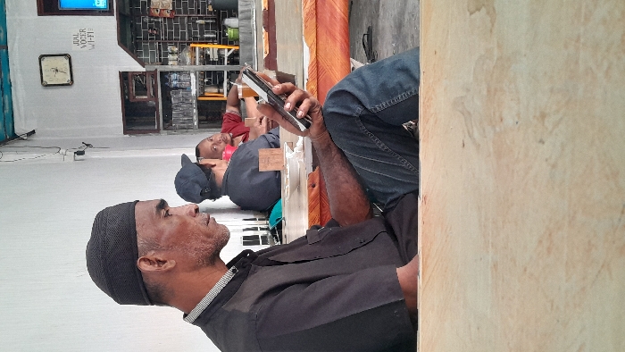 Three men are sitting down, seemingly on a ledge. The man closest to the camera is wearing a black cap and shirt. The man in the middle is wearing a black baseball cap and grey shirt. The man furthest away is wearing a red shirt. The man closest to the camera is holding a cell phone. The background is a white wall with a window. There is also a clock and text on the wall.
