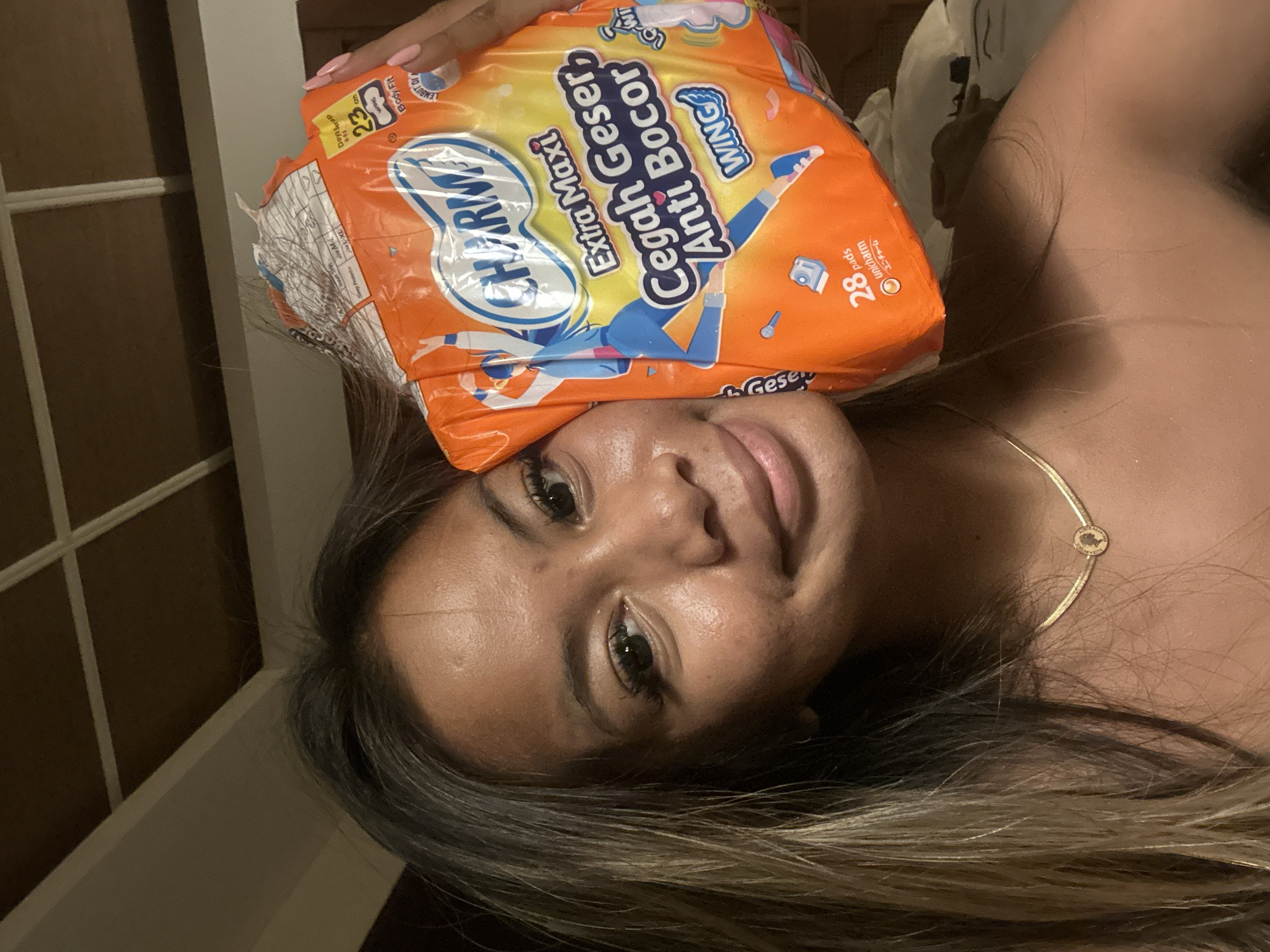A person with long, highlighted hair is holding a package of Charmee Extra Maxi Cegah Geser Anti Bocor sanitary pads on top of their head. The packaging is orange and blue, and it indicates that the pads have wings, are 23 cm in length, and the package contains 28 pads. The person is wearing a gold necklace with a circular pendant. A window is visible in the background. Based on the writing on the packaging, the image appears to be taken in Indonesia, however, there is no visible information in the image to confirm a specific city or district.
