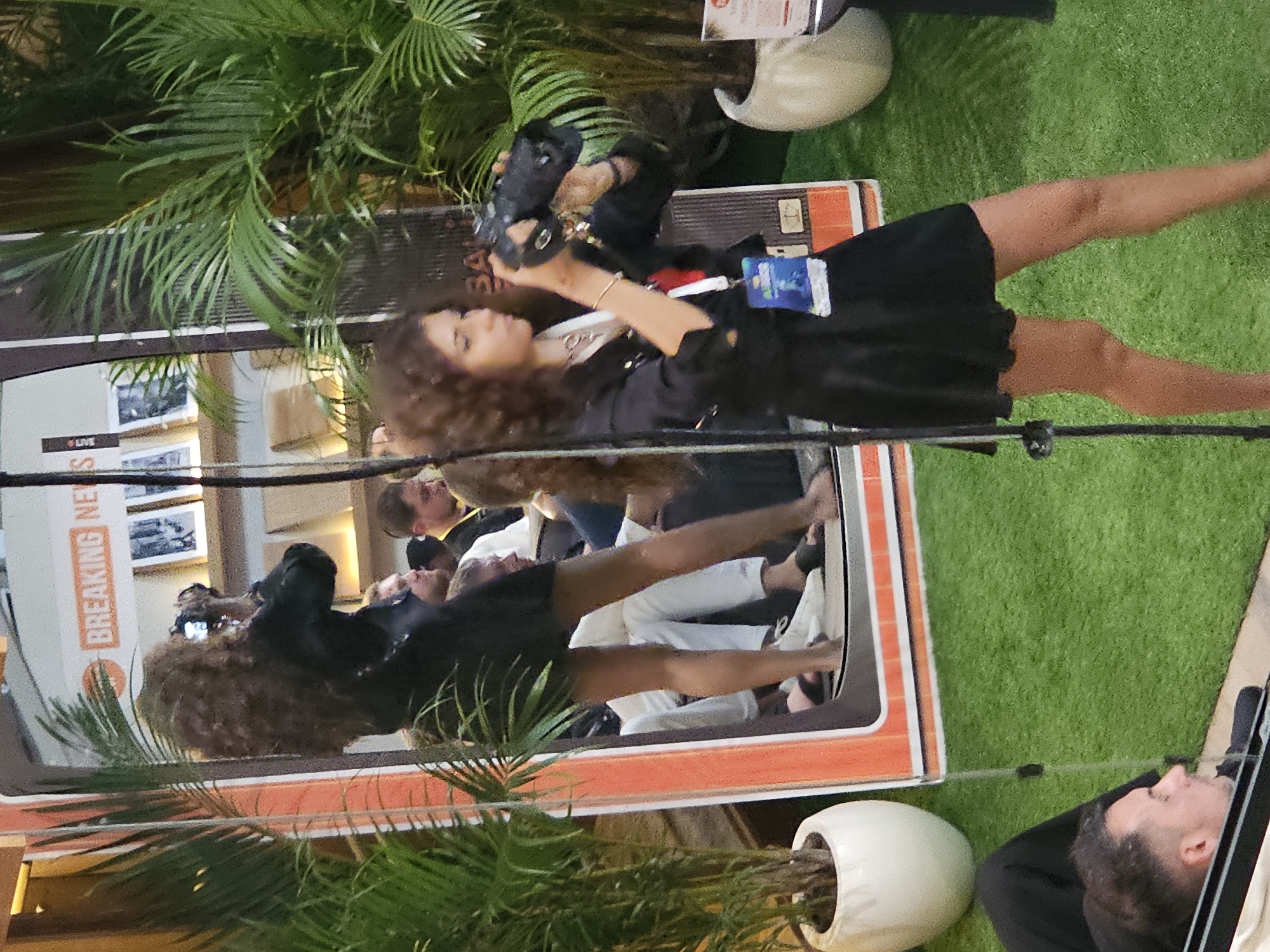 Captured at a possible promotional event in North Kuta, Indonesia, the image shows a group of people and objects, potentially related to the entertainment industry.
The most prominent figure is a woman with curly hair, wearing a black dress, holding a camera, and taking photos, possibly for media or event coverage. A crowd of other people appears in the background, with some facing forward.
The scene also includes potted palm trees, a sign with “BREAKING NEWS *LIVE” printed on it, and what looks like a photo booth or promotional display. The setting could be indoors.
