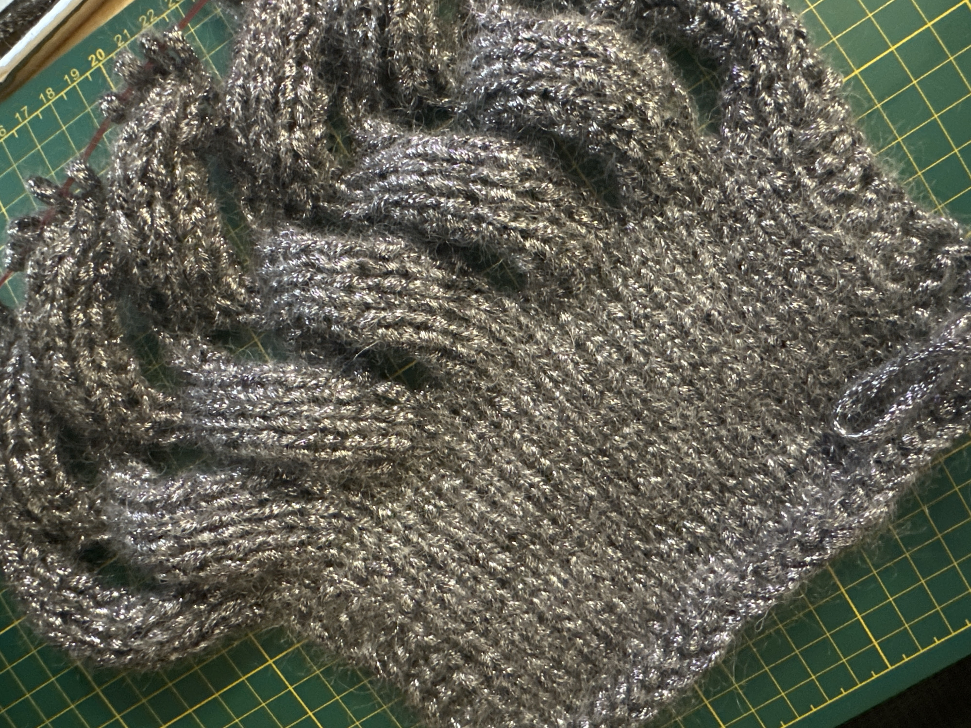 A close-up, overhead shot captures a grey, sparkly knitted fabric laid out on a green self-healing cutting mat with yellow grid lines. The fabric appears to be a portion of a garment or craft project, possibly a scarf or a sweater, with intricate knit patterns visible. The sparkly threads woven into the grey yarn catch the light, giving it a shimmering effect. The cutting mat's grid lines are partially visible beneath the fabric, suggesting the item is being worked on or displayed on a crafting surface. The background is out of focus, but hints of other materials or tools can be seen. There are no people or discernible environmental context beyond the crafting surface.