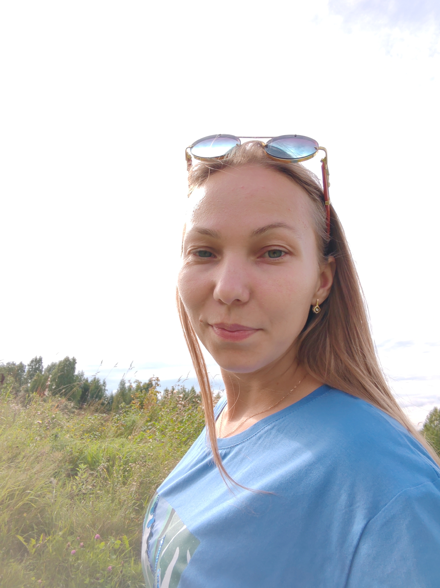 A young woman with long blonde hair is taking a selfie outdoors. She is wearing a bright blue t-shirt and sunglasses perched on her head. She has a delicate gold earring in her left ear and a thin gold chain around her neck. Her face is turned slightly towards the camera, and she is smiling subtly. The background shows a field of tall grass and trees under a bright, overcast sky. The location is implied to be within or near the city of Chukhlem, Russia, given the context provided.