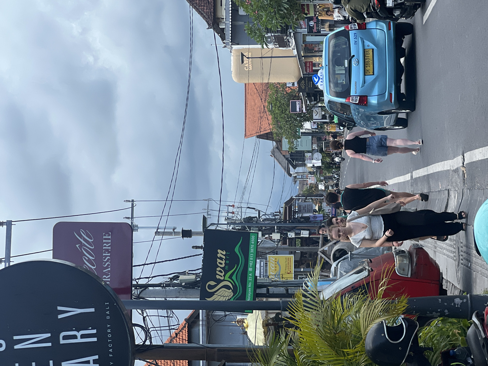 The image shows a street scene in Canggu, Indonesia. A light blue taxi with license plate "DK 1863 FBK" is driving down the street. Several pedestrians are walking on the side of the road. There are numerous shops and businesses visible, including "Swan mart," "ecote BRASSERIE," and "HENRY ARY FACTORY BALI." Power lines and poles are prominent. A red car is parked on the side of the road near a motorcycle with a black helmet. The sky is overcast.