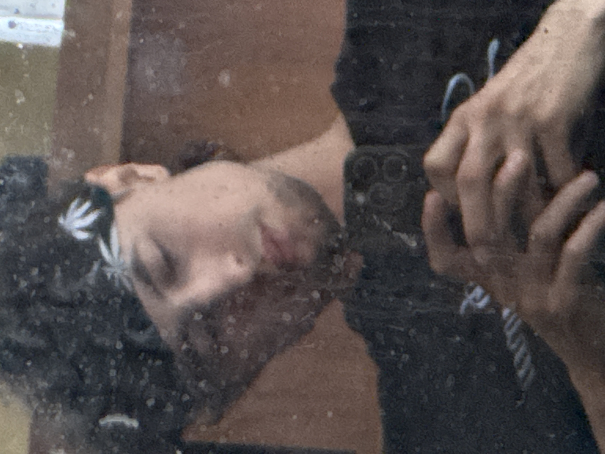 The image shows a close-up, slightly blurry, selfie-style photograph of a person's face reflected in a dirty mirror. The person is wearing a black bandana with a white leaf-like pattern and a black t-shirt. Their eyes are closed, and they are holding a smartphone. The background appears to be a wooden surface, possibly a shelf or cabinet. There are numerous small spots and smudges on the mirror's surface, obscuring some details of the reflection. The image does not contain any information about the location of Zaokskiy, Russia.