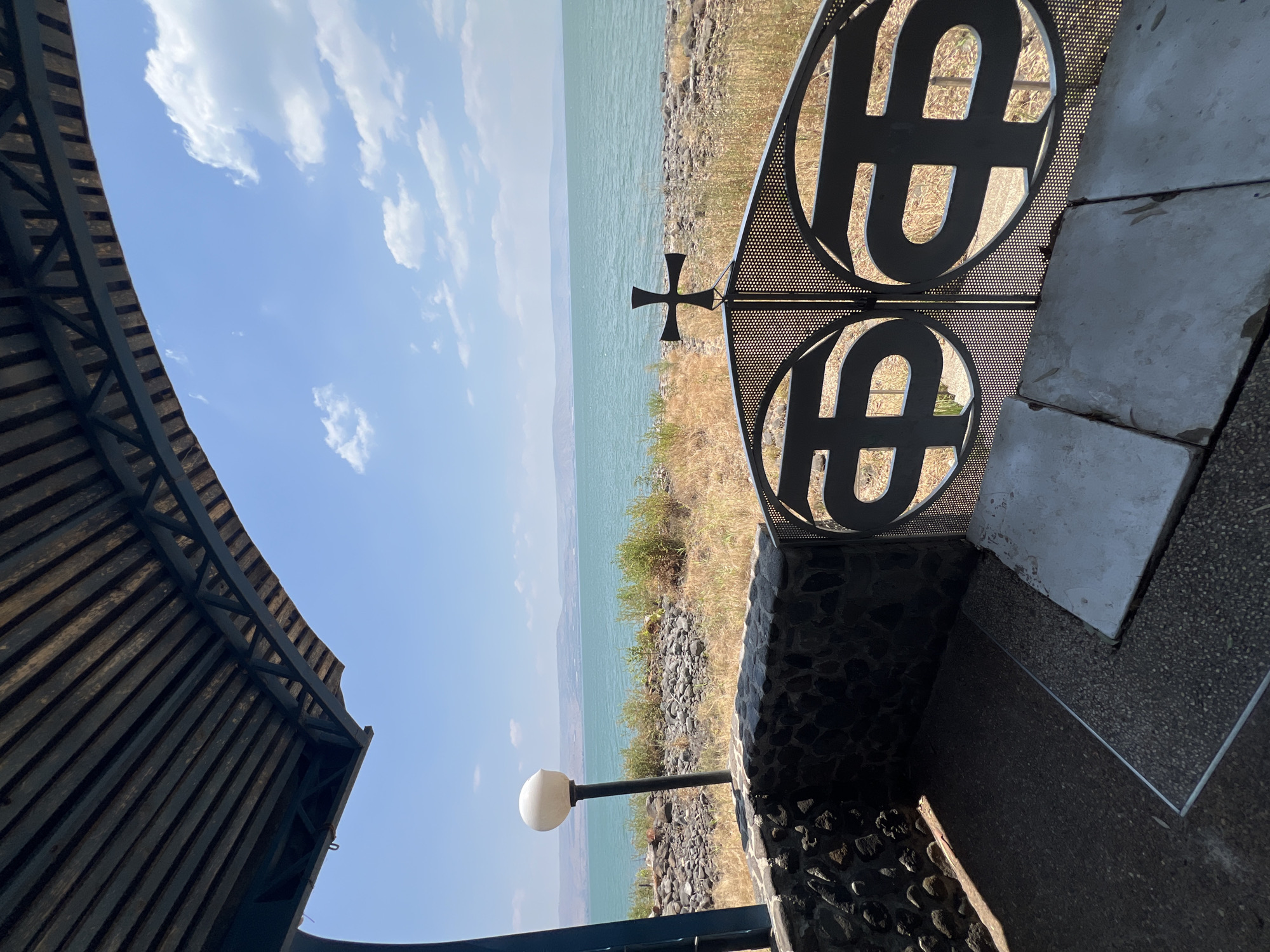The image captures a scenic view of the Sea of Galilee in Israel, from a sheltered outdoor area. The foreground features a stone wall and a wrought-iron gate with circular, abstract designs and a cross at its apex. A light fixture with a round, white globe stands to the left, near a textured stone pillar. The ground is paved with aggregate. Beyond the gate, the shore is lined with rocks and dry, golden vegetation. The Sea of Galilee stretches out in the mid-ground, its surface calm and turquoise under a clear blue sky with scattered white clouds. In the distance, rolling hills form the horizon. The overall impression is one of tranquility and natural beauty, suggesting a viewpoint overlooking the significant biblical site.