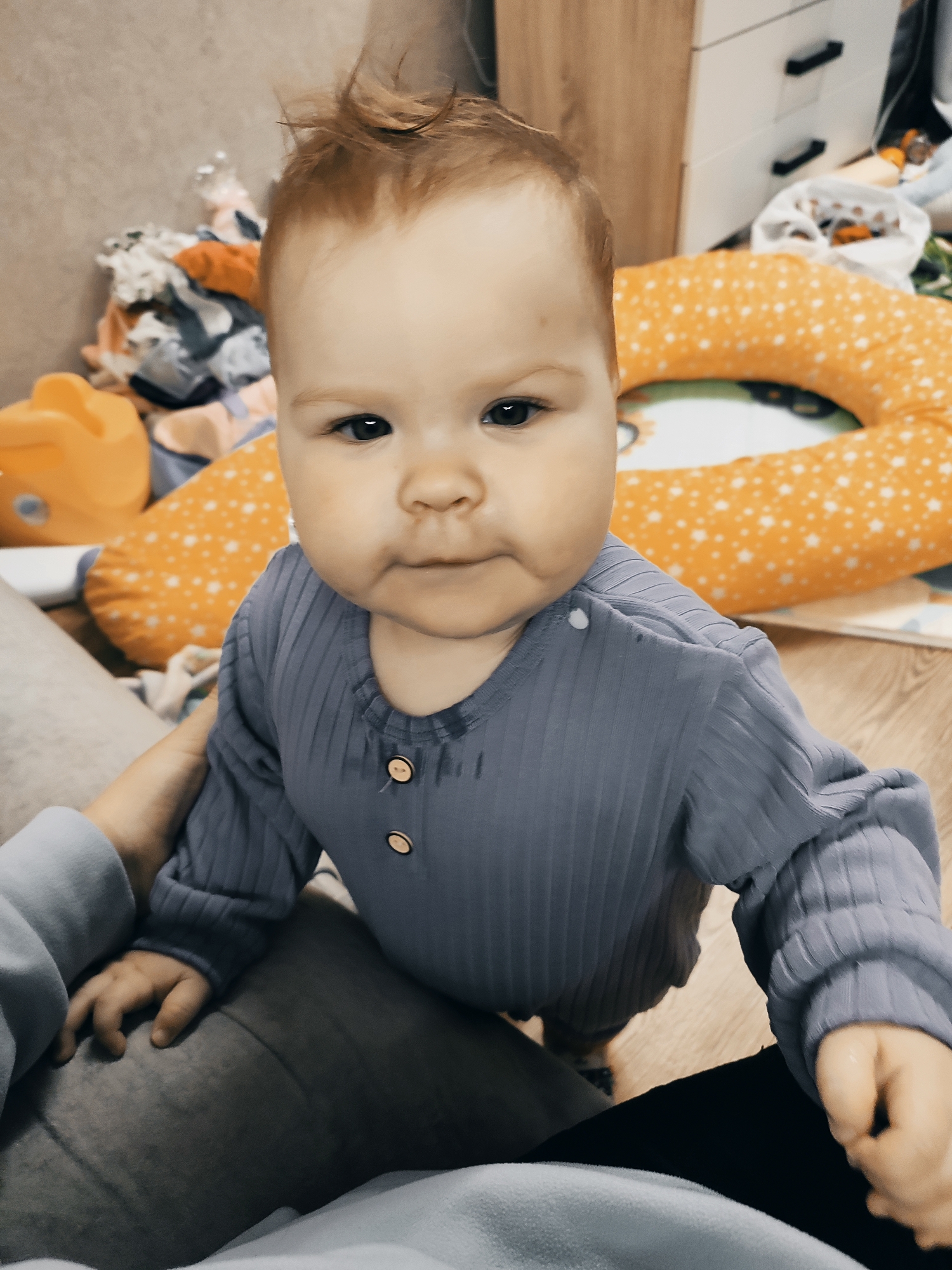 A baby with reddish-brown hair stands in front of the camera, wearing a long-sleeved, light blue ribbed shirt with two buttons at the neckline. The baby is being supported by an adult whose hand and portions of their clothing are visible on either side of the baby. The adult is wearing a light blue shirt and dark pants.
Behind the baby is a room setting containing an orange pillow with white stars, a pile of baby clothes, and a white cabinet in the background. The scene is indoors and appears to be in a home. There is a light-colored wooden floor. There is no information present to support the location being in the city of Chaikovsky, Russia.