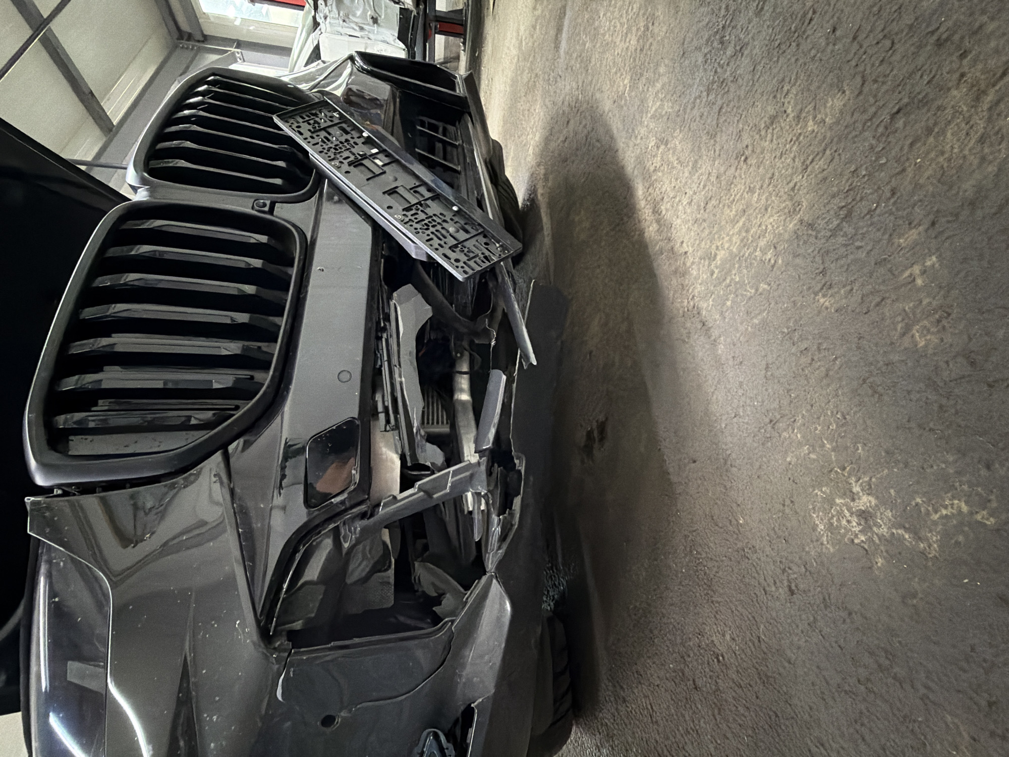 The image shows the front of a damaged black car, specifically the grille and front bumper area, which are significantly deformed. A license plate holder is detached and sits askew on the front of the vehicle. The car appears to be in a garage or repair shop, as evidenced by the concrete wall to the right and the partially visible interior of the space behind the car. There are no people visible. The location is unknown and no details confirm it being in Burgas, Bulgaria.