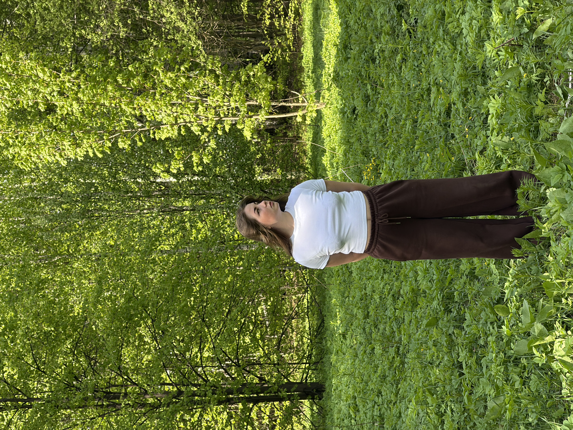 A young woman stands in a grassy area surrounded by trees with bright green foliage. She wears a white t-shirt and brown sweatpants. The location appears to be a park or wooded area, possibly in the vicinity of Ural'skoe, Russia, based on the assumption of geographic location. There are no observable interactions. The environment is bright and sunny.