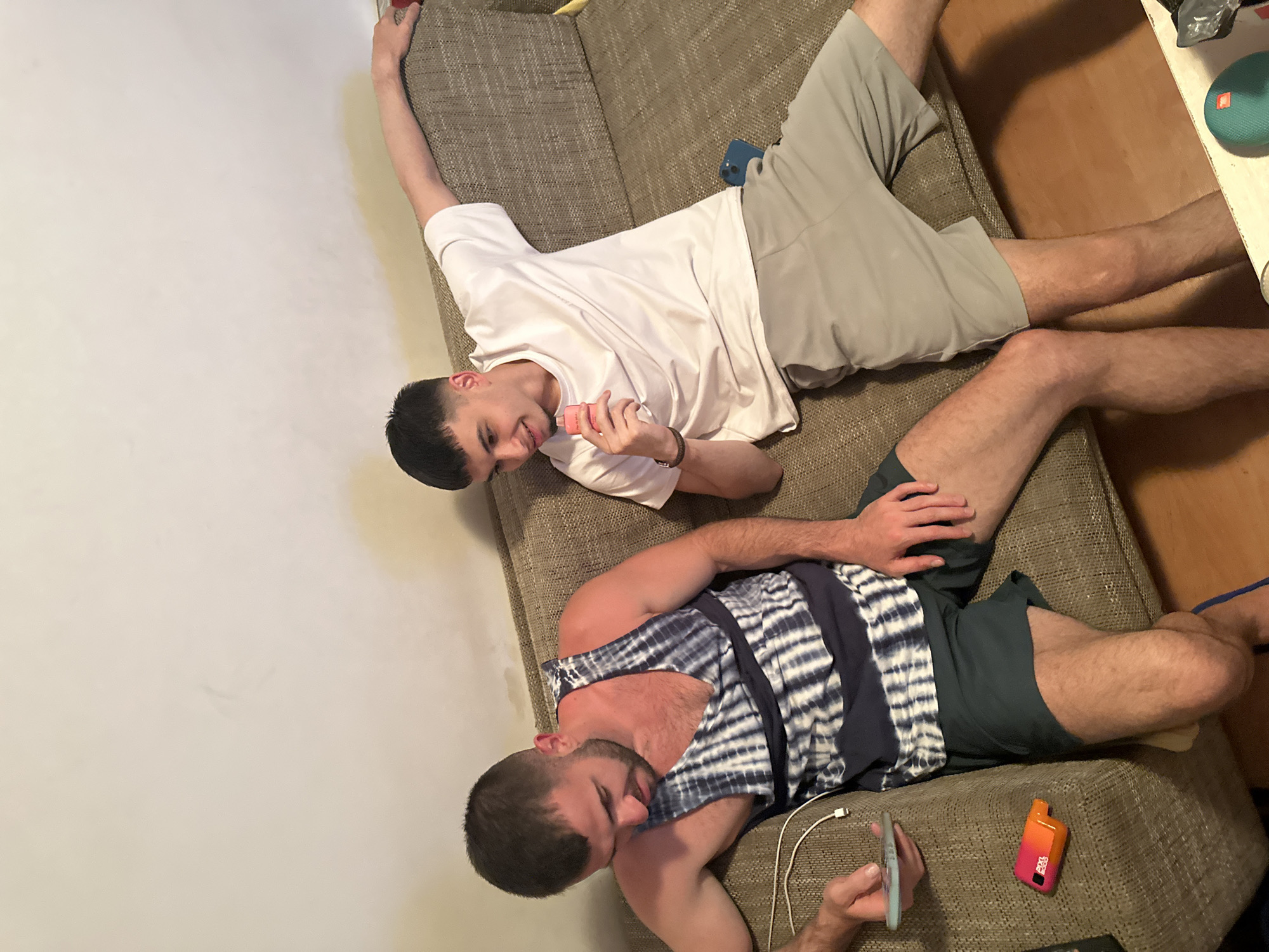 The image displays two young men relaxing on a brown fabric sofa in what appears to be an indoor setting, possibly a living room, in Sofia, Bulgaria. The room features a light-colored wall on the left and a wooden floor with a white side table on the right.
The man positioned higher on the sofa is lying on his back, dressed in a white short-sleeved t-shirt and light grey shorts. He has short dark hair with a fade and a gentle smile, holding a small pink object, possibly a vape or a cosmetic item, in his right hand. His left arm is casually stretched upwards, and a dark blue smartphone is partially visible beneath his leg.
The second man is lower on the sofa, lying on his right side. He is wearing a blue and white tie-dye tank top and dark grey shorts, with short dark hair and a beard. He is engrossed in a light blue smartphone held in his left hand, which is connected to a white charging cable. An orange and pink vape device, clearly labeled "PULSE X BAR," rests on the sofa beside his right hand.
On the white side table to the right, a teal JBL portable speaker displaying its "JBL" logo is visible, along with a black plastic bag or item. Both individuals appear relaxed and absorbed in their personal devices, indicating a casual and leisurely atmosphere. The lighting suggests it is likely daytime indoors.