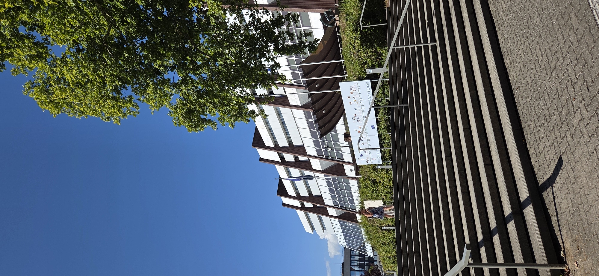 A wide-angle, low-angle shot captures the exterior of a modern building in Strasbourg, France, under a clear blue sky. The building, characterized by its white facade with dark brown angular accents and numerous windows, rises behind a lush green hedge. In front of the building, a large informational sign displays various flags and text. A woman in a floral dress stands on a wide, grey stone staircase leading up to the building, looking to her right. The staircase is bordered by metal railings and casts shadows on the patterned pavement below. A mature tree with vibrant green leaves partially obscures the top right portion of the building and the sky.