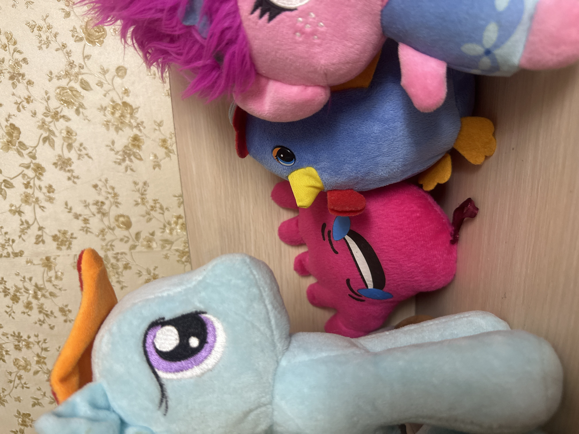A collection of stuffed toys is arranged on a wooden shelf against a backdrop of floral wallpaper. In the foreground, a light blue plush toy with orange ears and a purple eye is positioned on the left. To its right, a bright pink plush toy with a smiling face is lying down. Next to it is a blue plush toy resembling a bird with a yellow beak and red comb. Partially visible on the far right is a pink plush toy with bright pink hair. The toys are positioned closely together, creating a playful scene. The setting does not provide any specific clues to determine its location within Kazan, Russia.