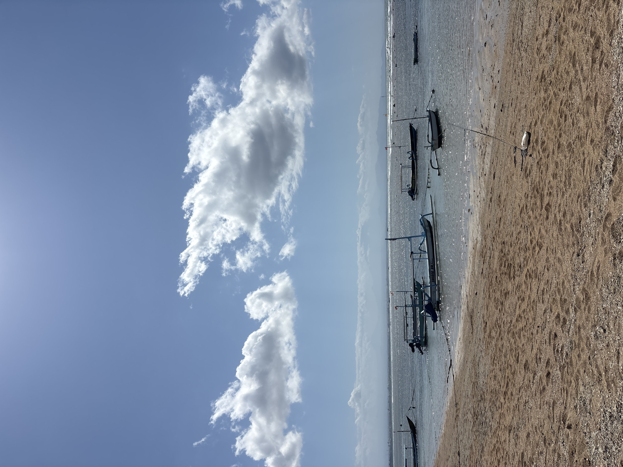 The image depicts a tranquil beach scene in Denpasar, Indonesia, under a clear blue sky dotted with fluffy white clouds. The foreground is dominated by a sandy beach, its surface a mix of fine sand and small pebbles, with a rope and buoy anchoring a small boat in the shallow water.
The water itself is calm, with gentle waves lapping at the shore. Several traditional Balinese fishing boats, known as "jukung," are moored in the water, their slender hulls and outriggers characteristic of the region. These boats vary in color, with some featuring blue accents, and one clearly visible boat bears the inscription "SAHARA BALI." The boats are anchored at various distances from the shore, some appearing closer to the beach while others are further out, creating a sense of depth.
In the distance, the horizon is marked by a hazy, mountainous landscape, suggesting the presence of land beyond the immediate coastal area. The overall atmosphere is one of serenity and peacefulness, characteristic of a tropical beach setting. The bright sunlight casts a gentle sheen on the water, highlighting the textures of the sand and the boats. The scene is devoid of people, emphasizing the natural beauty of the location.