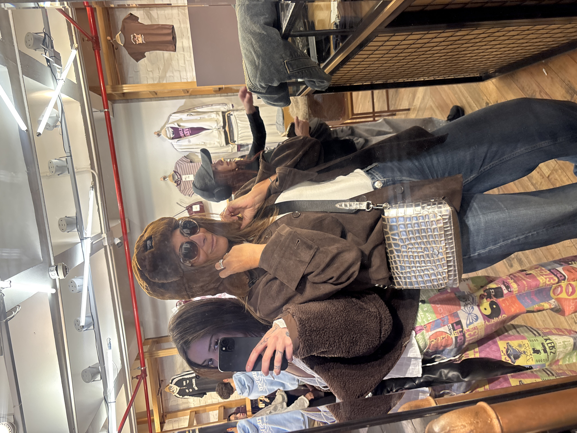 A woman is taking a mirror selfie in a store. The image appears to be a reflection, as evidenced by the orientation of the subject holding the phone. The woman is wearing a brown furry hat and sunglasses. Another person is visible behind her in the mirror. Clothes are hanging on racks in the background. The reflection in the mirror creates the illusion of multiple layers of clothing and items. The exact location in Buenos Aires, Argentina, cannot be determined from the image alone, as there are no distinct landmarks visible.