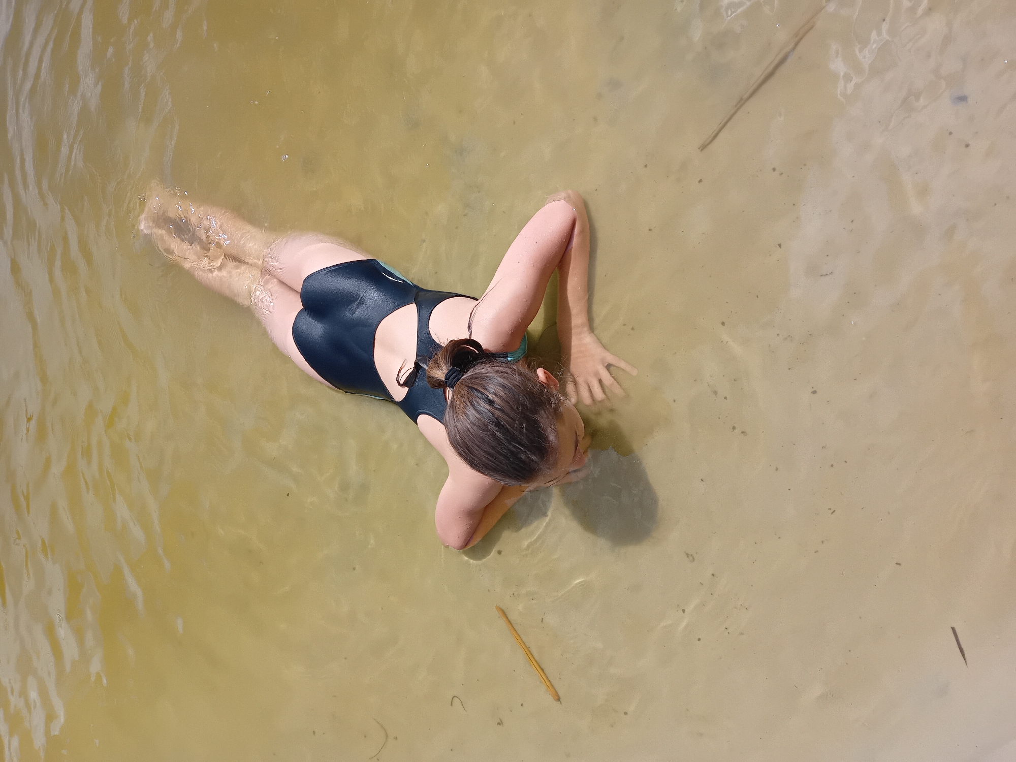 In the city of Chuhuiv, Ukraine, a person in a black swimsuit is partially submerged in murky, shallow water, possibly a river or lake, based on the sandy bottom visible beneath the surface. The individual is lying on their front, facing downwards towards the water, with their head tilted to the side and resting on their arms. Their legs are extended behind them, visible just below the surface. Two thin, dry reeds or sticks lie in the water near the person. The water is yellowish-brown and appears slightly cloudy, with ripples and reflections indicating movement from an unseen source. The overall scene suggests a natural, outdoor environment.