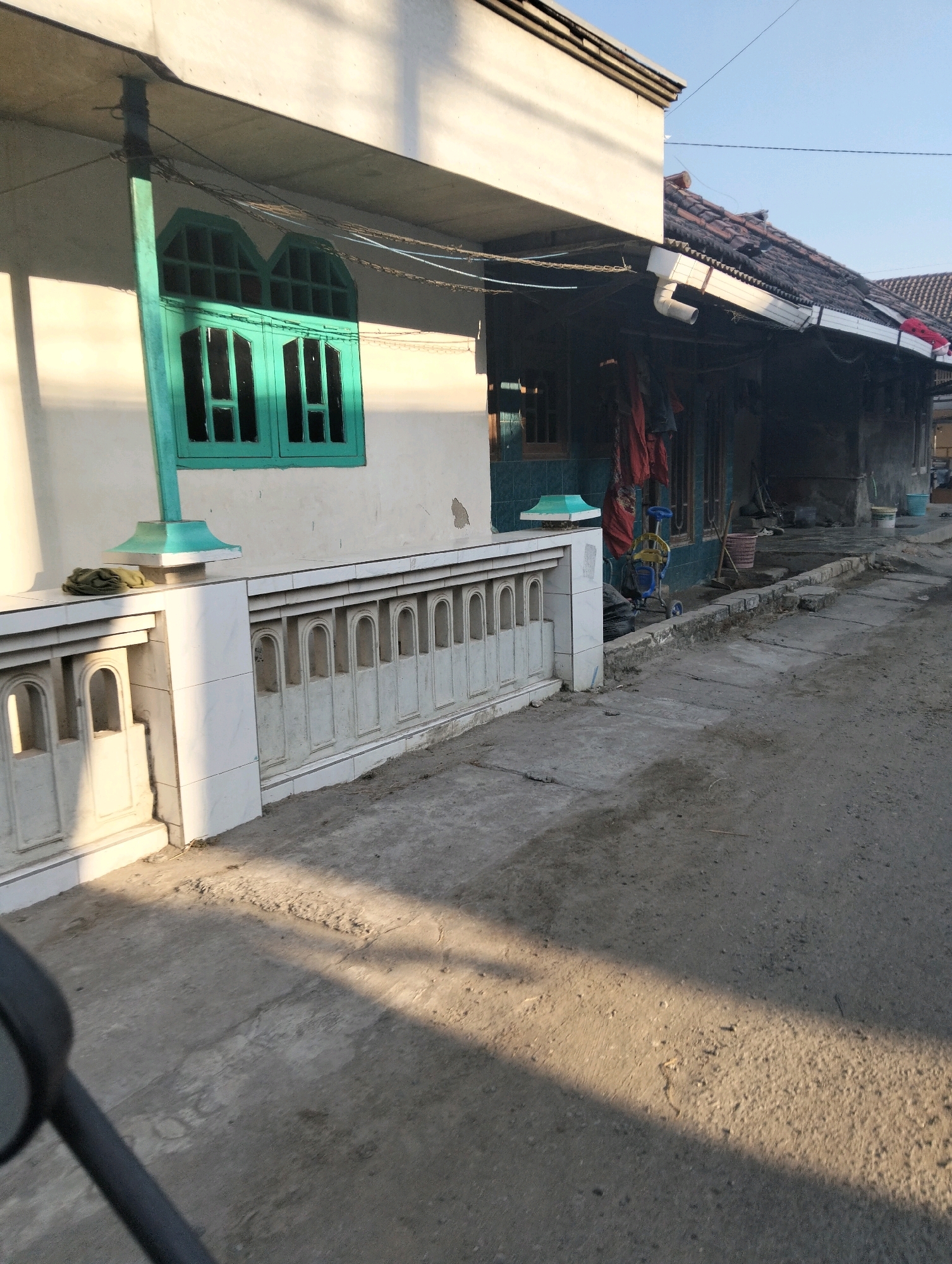 The image depicts a street scene in Drajat, Indonesia, under a clear blue sky. Sunlight casts long shadows across a concrete path and the front of a building. The building's facade is painted white, with a distinctive turquoise window frame with arched top sections. Adjacent to the window is a white tiled balustrade. To the right, the image shows a row of structures with tiled roofs, some appearing weathered. In front of these structures, there is a collection of items including red cloth draped, a small blue and yellow stroller, a basket, a broom, and buckets. The ground is predominantly concrete and dirt, showing signs of wear and tear. Part of a dark grey object, likely a motorcycle mirror, is visible in the lower left corner.