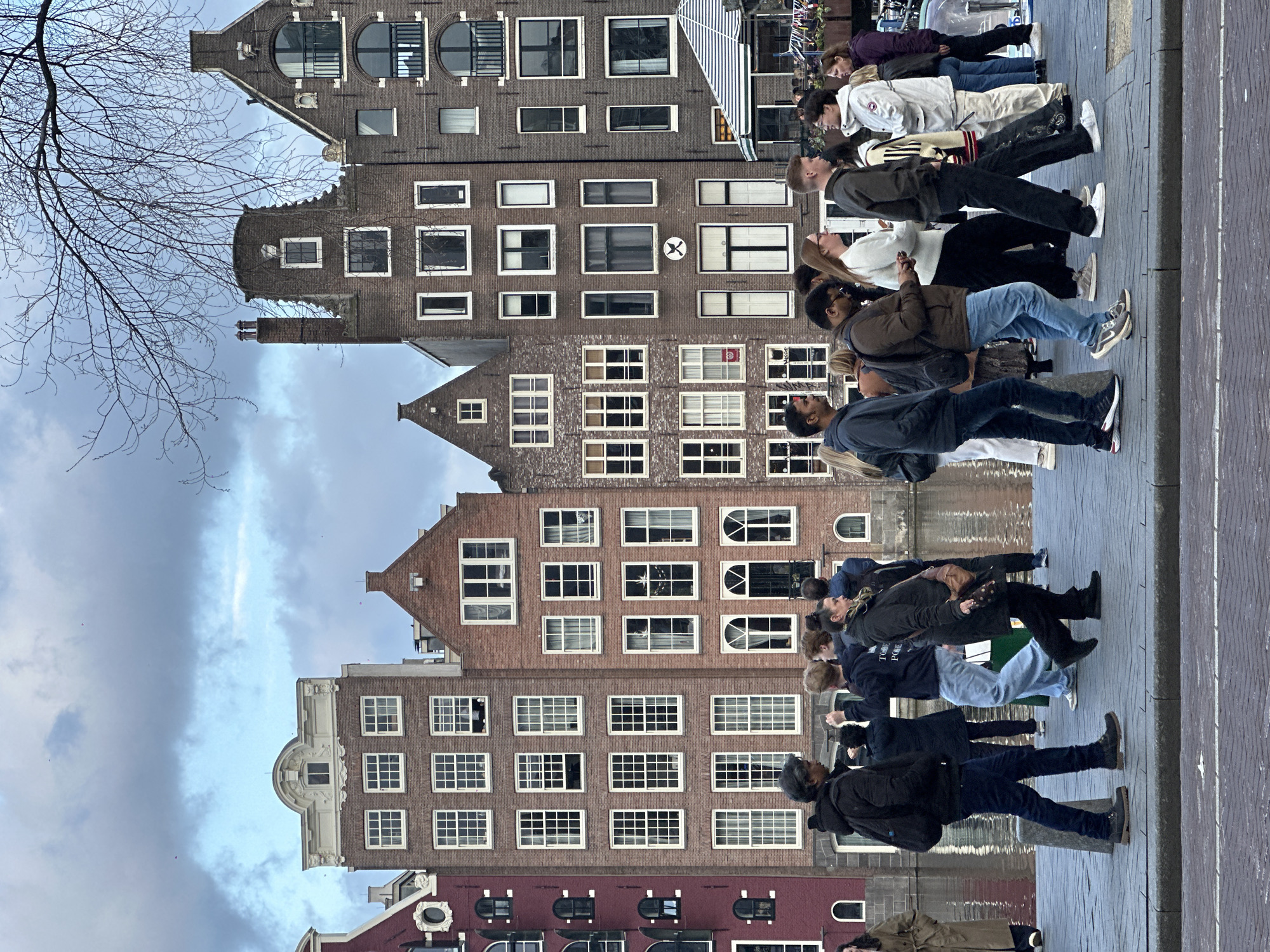 The photograph shows a group of people lined up along a canal in Amsterdam, Netherlands, likely admiring or taking photos of the historic canal houses. The architecture of the buildings is characterized by their narrow facades, steep gables, and numerous windows. The buildings are constructed of brick in various shades of red and brown. Some have decorative elements such as carved stone details. Bare branches of a tree are visible in the upper left corner. The sky is overcast with a mix of gray and white clouds. The pavement along the canal is brick. The people are wearing casual clothing and appear to be tourists.
