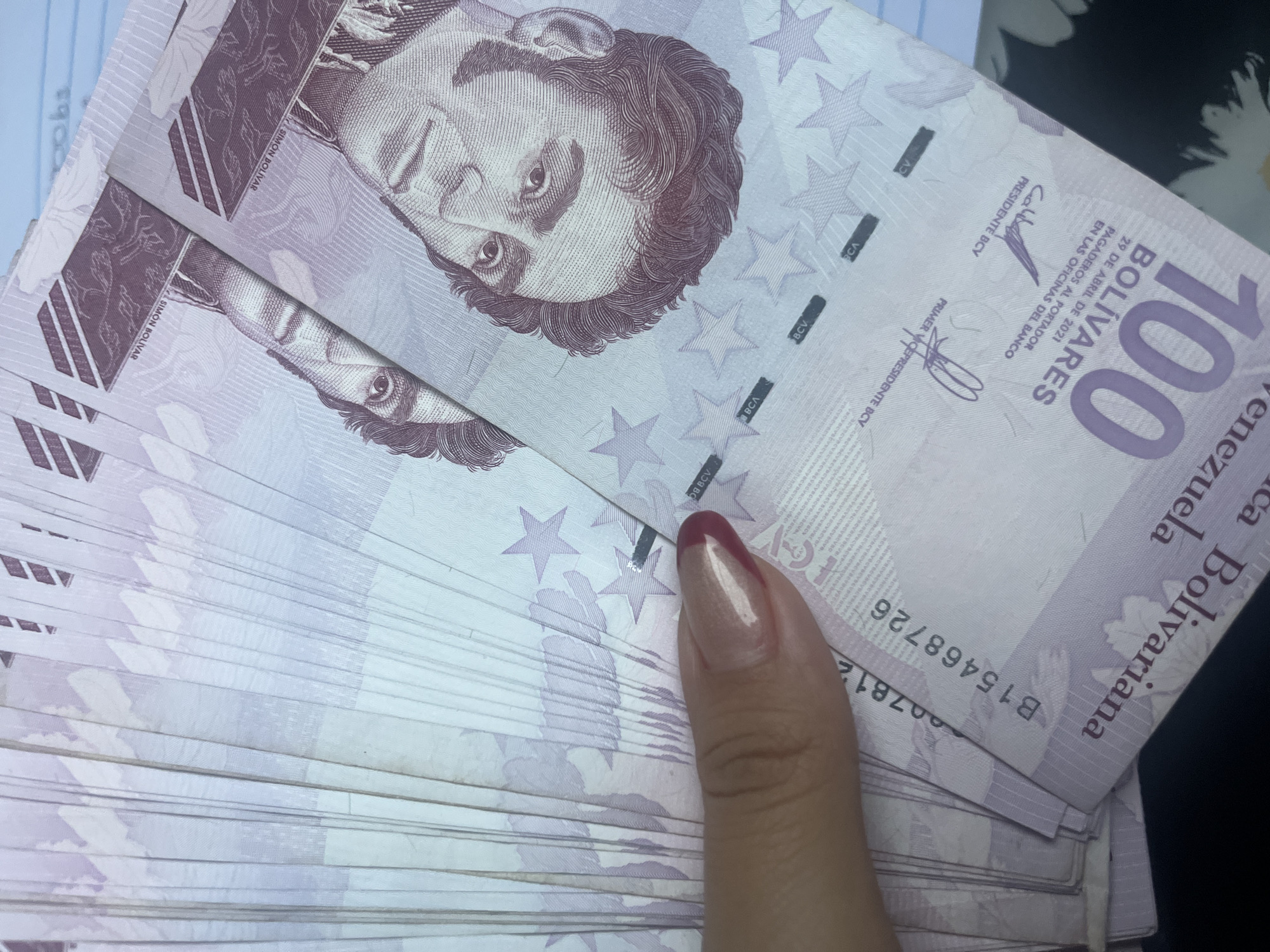 A close-up, overhead shot displays a fan of Venezuelan 100 Bolivares banknotes. The denomination "100 BOLIVARES" is prominently visible on the lower right side of the bills. The portrait of Simón Bolívar appears on the upper right portion of the notes. A woman's hand, with a pinkish-gold colored nail polish and a dark red accent on the tip of one nail, is holding the banknotes. The background is blurred, but appears to be a flat surface. There is no indication of the specific location within Conuco Viejo, Venezuela.