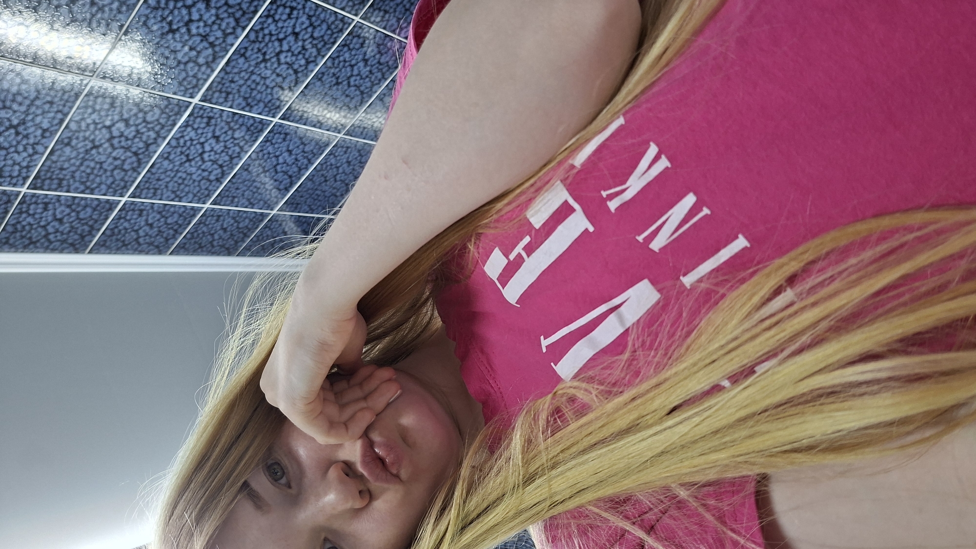 A young woman with long blonde hair is pictured indoors. She is wearing a pink t-shirt with white lettering that includes the word "INK." She is looking at the camera, making a kissing face, and holding her left hand in front of her face, partially covering her left eye. The background consists of an institutional tile ceiling with a speckled blue pattern and a grey wall panel. The exact location in Chaikovsky, Russia, cannot be determined from the image.