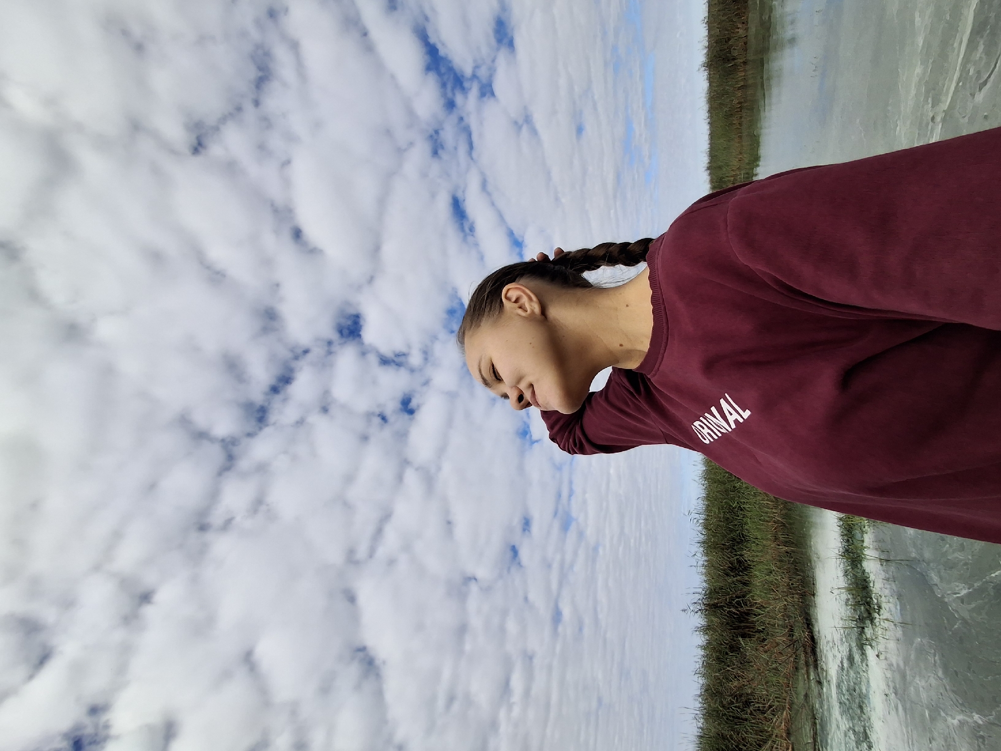 In Plishkino, Russia, a young woman with braided brown hair is captured from the chest up, her face turned upward with her eyes closed in a moment of peaceful contemplation. She wears a dark red sweatshirt with white lettering that reads "ORINAL" across the chest. Her right arm is raised, with her hand resting on the back of her head. The background is dominated by a vast expanse of sky filled with white, puffy clouds, against which a vibrant blue sky is visible in patches. Below the sky, a calm body of water, tinged with a greenish hue, reflects the clouds. A dense line of tall, green reeds borders the water. The overall scene suggests an outdoor setting, possibly a lake or riverbank, under a cloudy but bright sky.