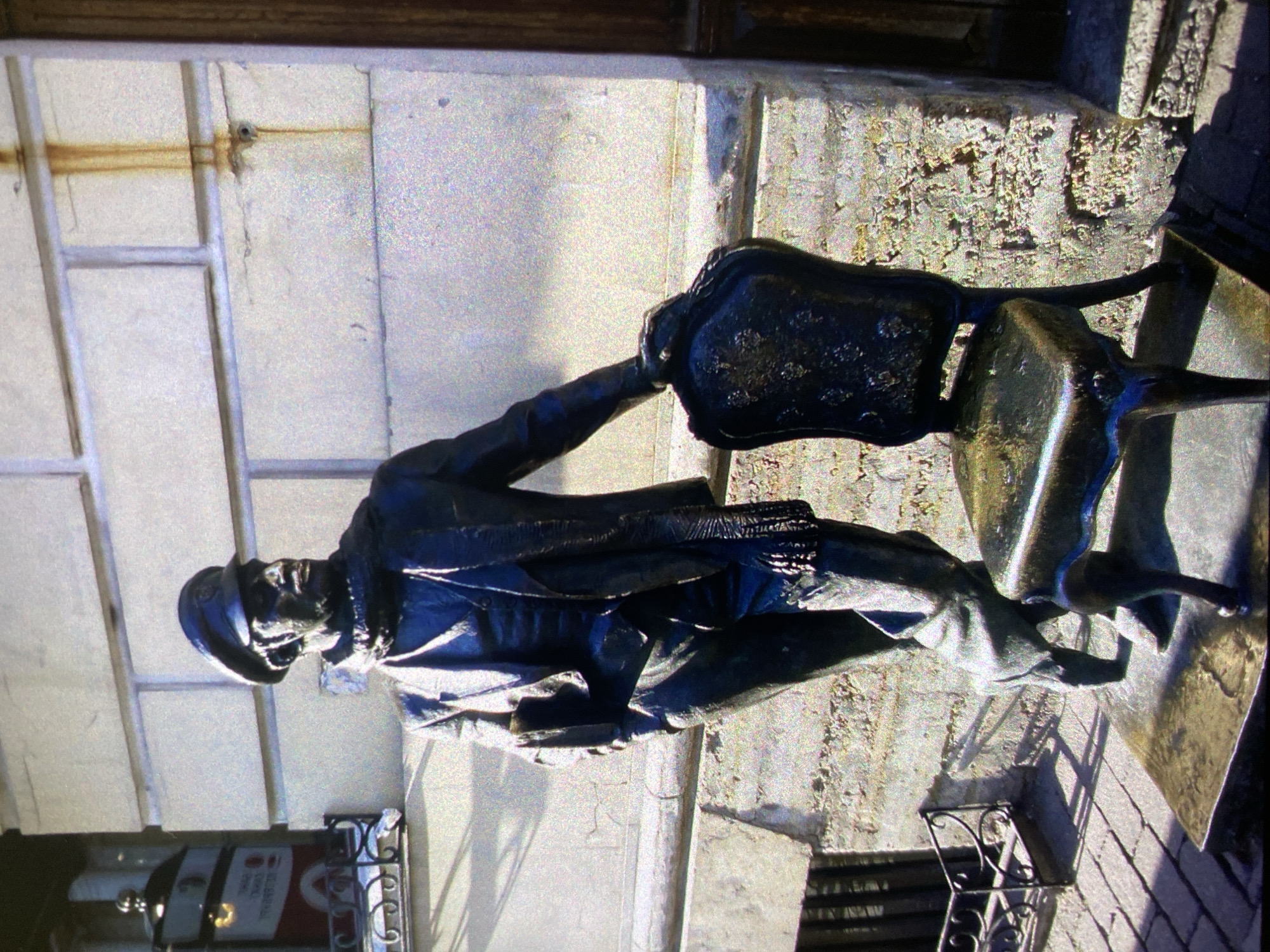 A medium shot depicts a bronze statue of a seated figure in Saint Petersburg, Russia. The figure, wearing a hat and a formal coat, is grasping the back of an ornate metal chair. The statue is positioned on a weathered, textured surface. The background consists of a white wall with a metal grid-like structure, and signs in foreign language below. The overall lighting is bright, giving the statue a polished appearance.