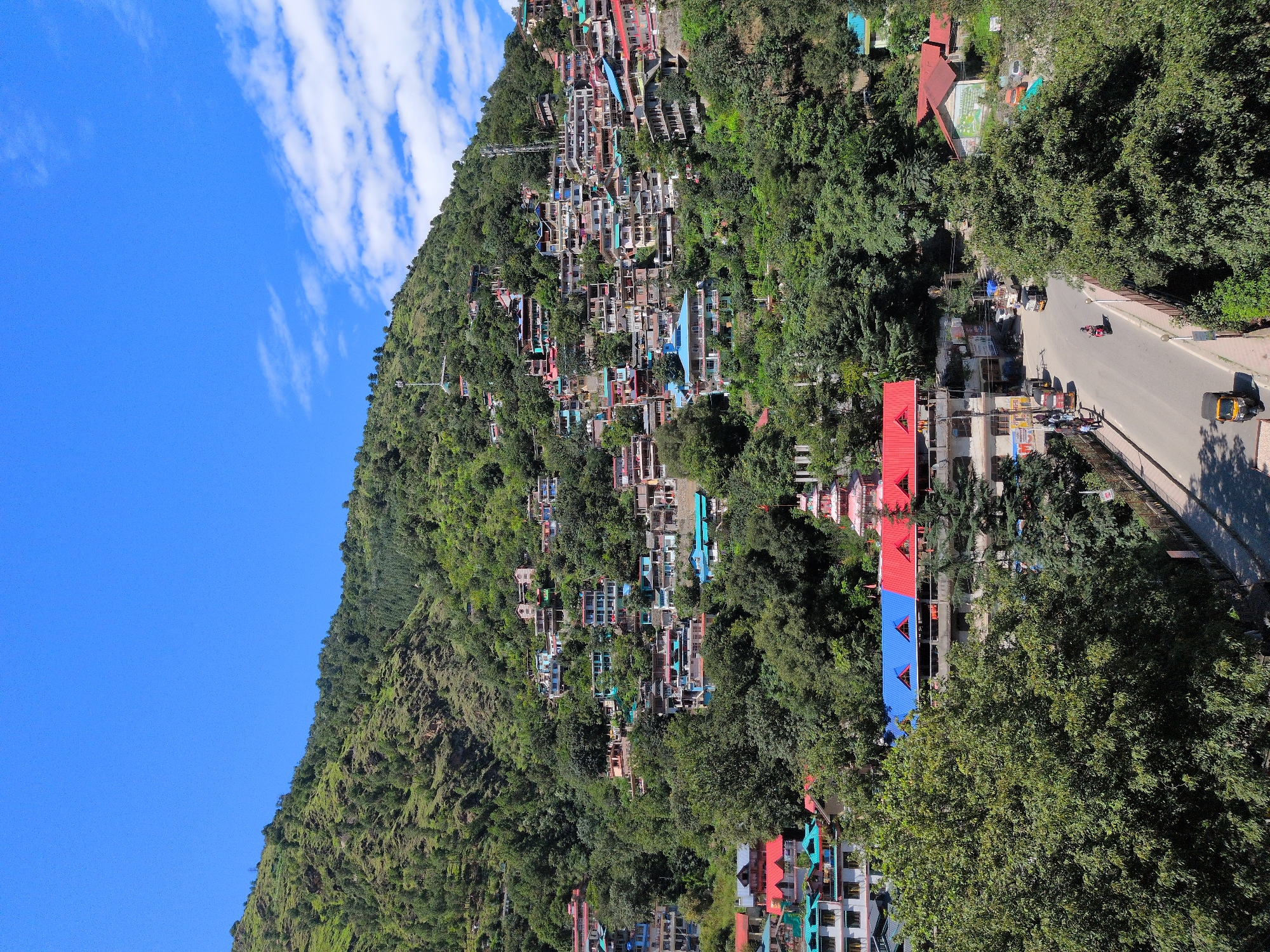 An elevated, high-angle, medium shot captures a hillside town in Kullu, India, under a clear blue sky with scattered white clouds. The town sprawls across a densely forested green mountain, with buildings of various sizes and colors clustered together. Many structures feature red and blue roofs, some with balconies. Vegetation is abundant, with trees and shrubs interspersed among the buildings and blanketing the mountain slopes. A road curves through the foreground, with several vehicles, including a scooter, auto-rickshaws, and a truck, in motion or parked. Pedestrians are visible on the sidewalks. A prominent temple-like structure with tiered red roofs stands out amidst the buildings. The scene depicts a bustling community integrated with its natural mountainous environment.