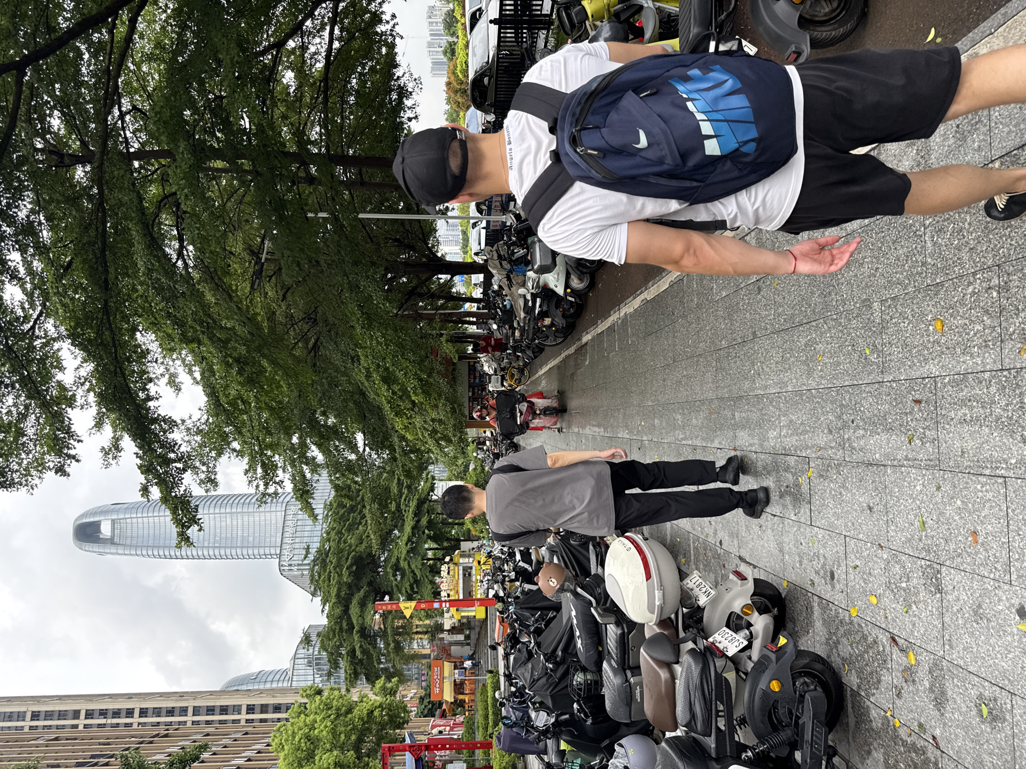 In Haizhu District, China, on a cloudy day, two men walk down a wet, paved sidewalk lined with numerous parked electric scooters and motorcycles. The man in the foreground, facing away from the camera, wears a white t-shirt, black shorts, a black baseball cap, and a dark blue backpack with a Nike logo. He has a red bracelet on his left wrist. To his left, another man walks in the same direction, facing away from the camera, wearing a gray t-shirt and black pants. Behind him, other individuals are visible, including one riding a scooter. The sidewalk is bordered by a row of lush green trees on the right and a line of parked scooters on the left, some covered with black tarps. In the background, tall buildings, including a distinct skyscraper with a curved top, and more trees can be seen. Cars are parked on the street to the right, and what appear to be commercial establishments are visible further down the sidewalk on the left. Fallen yellow leaves are scattered on the wet pavement.