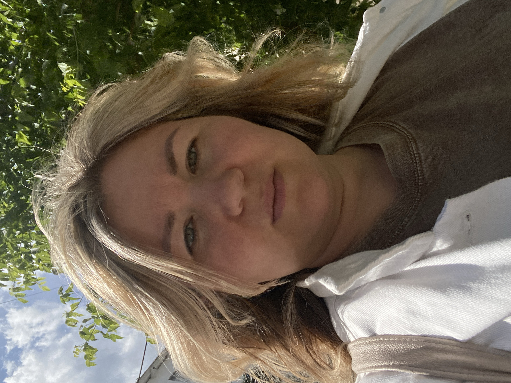 A woman with blonde hair and green eyes is taking a selfie outdoors. She is wearing a white denim jacket over a brown t-shirt. A light-colored strap from a bag is visible over her left shoulder. She is standing in front of a backdrop of lush green trees and a blue sky with white clouds. The lighting suggests it is daytime. The location is identified as Chaikovsky, Russia.