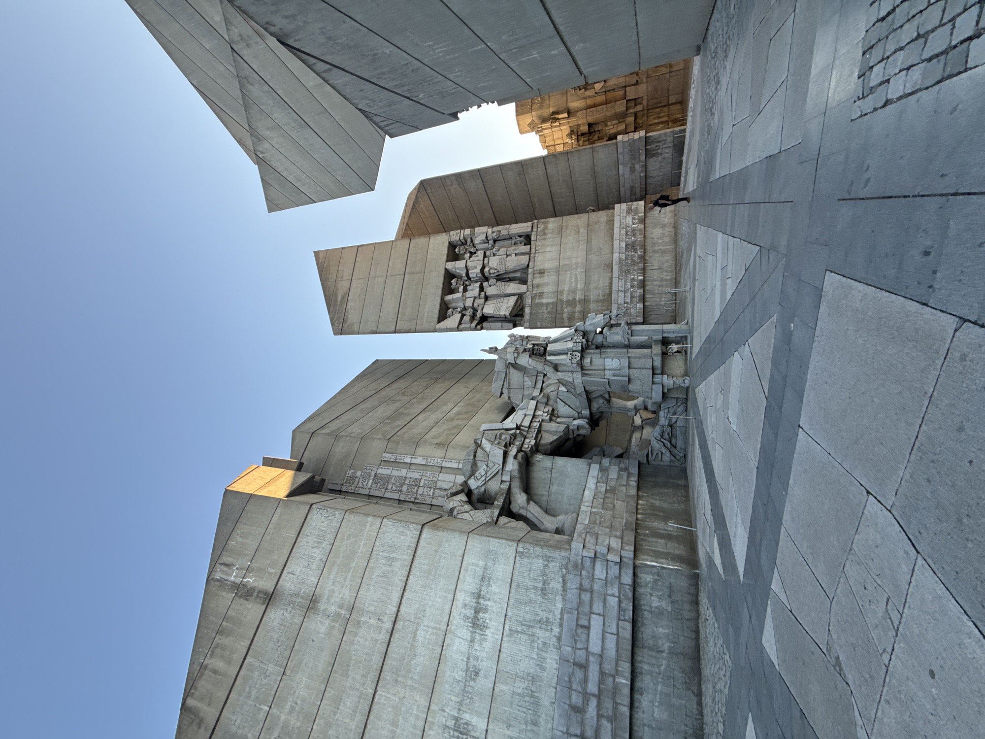 The image features the Founders of the Bulgarian State Monument in Shumen, Bulgaria. The monument is composed of large, angular concrete blocks forming a complex geometric structure. These blocks are adorned with sculptures and inscriptions. One block has a prominent relief depicting figures, seemingly historical figures. Below this, a sculptural composition is visible. The ground is paved with large, gray stone slabs, creating a pattern that is visible from the low angle of the shot. In the background, a person is walking away from the viewer, adding a sense of scale to the structure. The sky is visible between the blocks, it is blue and clear.