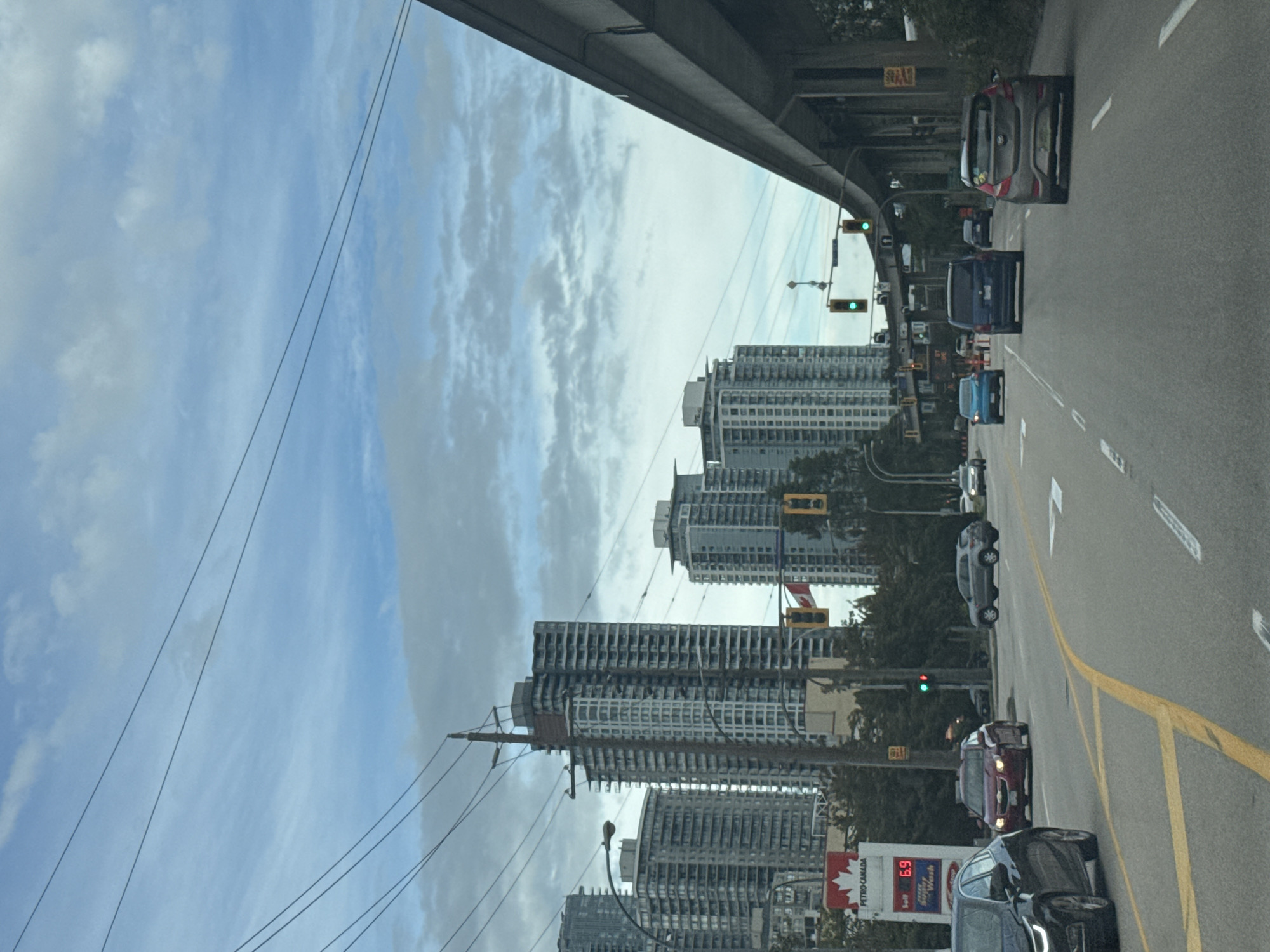 The image shows a street scene in New Westminster, Canada, on a cloudy day. The foreground features a multi-lane road with several cars driving in the same direction. The middle ground is dominated by tall apartment buildings and a Petro-Canada gas station with a price of 6.9 displayed on its sign. In the background, more skyscrapers and an elevated highway structure are visible. The sky is partly cloudy, with streaks of white clouds against a pale blue background. There are power lines stretching across the frame. The image appears to be taken from inside a moving vehicle, indicated by the perspective and the blurred motion of some elements.