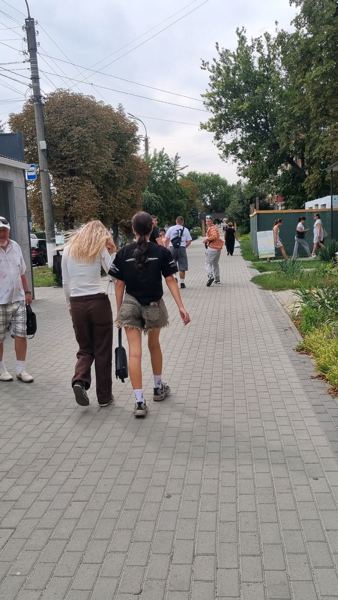 A group of people walk along a paved sidewalk in Smila, Ukraine. The sky is overcast. On the left, a man in a white shirt and plaid shorts walks away from the camera. In the center, two young women walk away from the camera. The woman on the left has blonde hair and wears a white long-sleeved shirt and brown pants. The woman on the right has dark hair in a ponytail and wears a black t-shirt and denim shorts. Other people are visible in the background walking on the sidewalk or standing. Trees and buildings line the sidewalk.