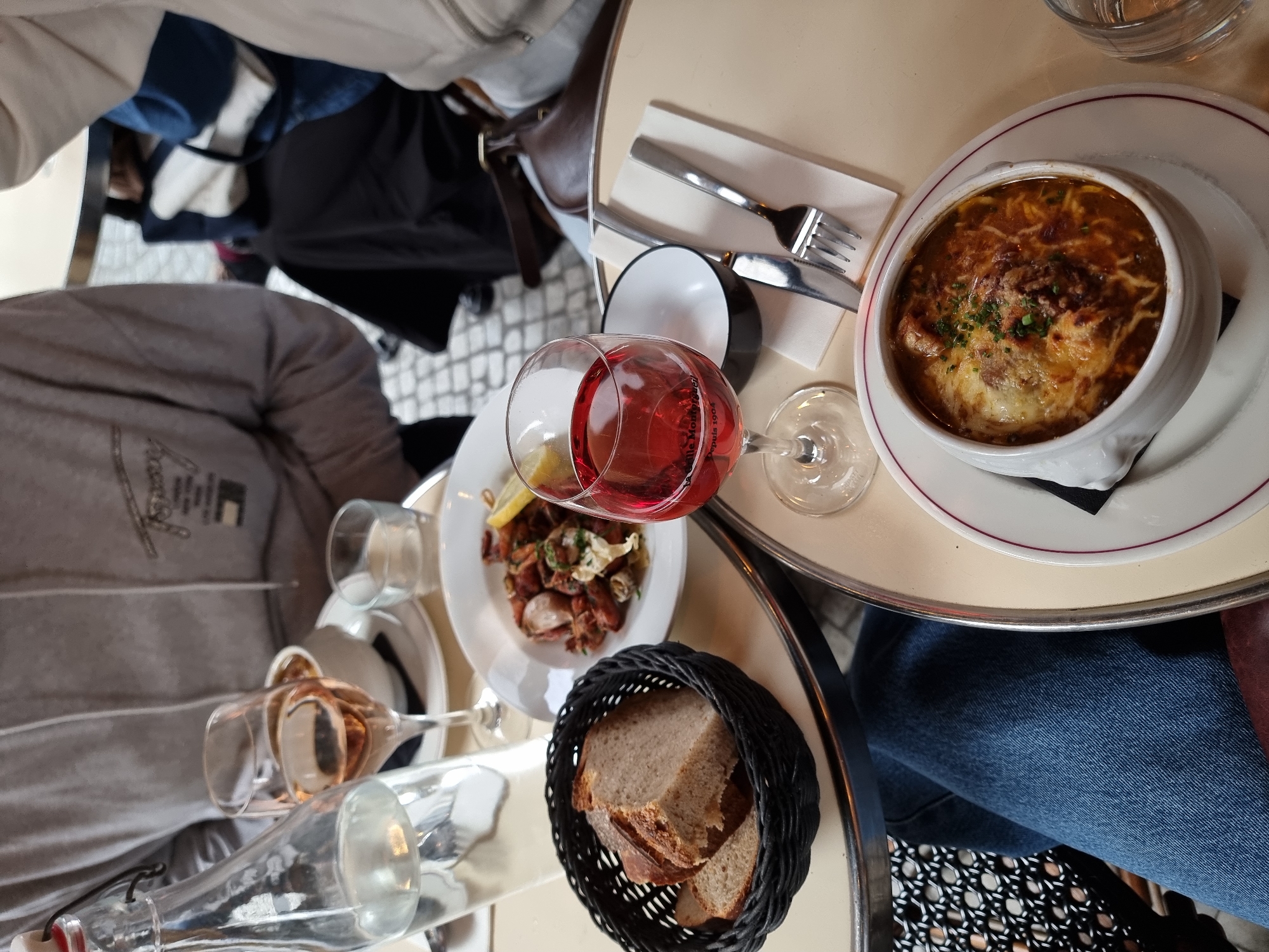 The image shows an overhead view of a table at a Parisian cafe. The table is circular with a beige top and a metal rim. A person wearing a gray sweatshirt is partially visible on the left side of the image, while another person wearing jeans is on the right.
On the table, there is a bowl of French onion soup served on a white plate with a red rim, silverware, a glass of rosé wine, a small plate of food with a lemon wedge, and a basket of bread. There are also several empty glasses and a bottle of water.
The background shows other patrons of the cafe, bags, and clothing. The ground is visible and patterned with white and gray tiles. The location can be confidently identified as a Parisian cafe based on the classic bistro table and chair design, the presence of traditional French food items, and the general ambiance.