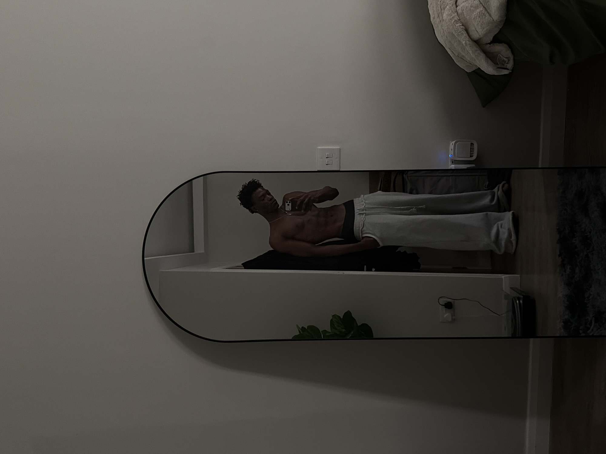 A photograph shows a shirtless man in a mirror wearing gray sweatpants. The man is holding his phone to take a picture of himself. The background shows a white wall, a white shelf, a bed, a white and green blanket, a white plug, and a houseplant. There is no evidence indicating this scene is in London, United Kingdom.