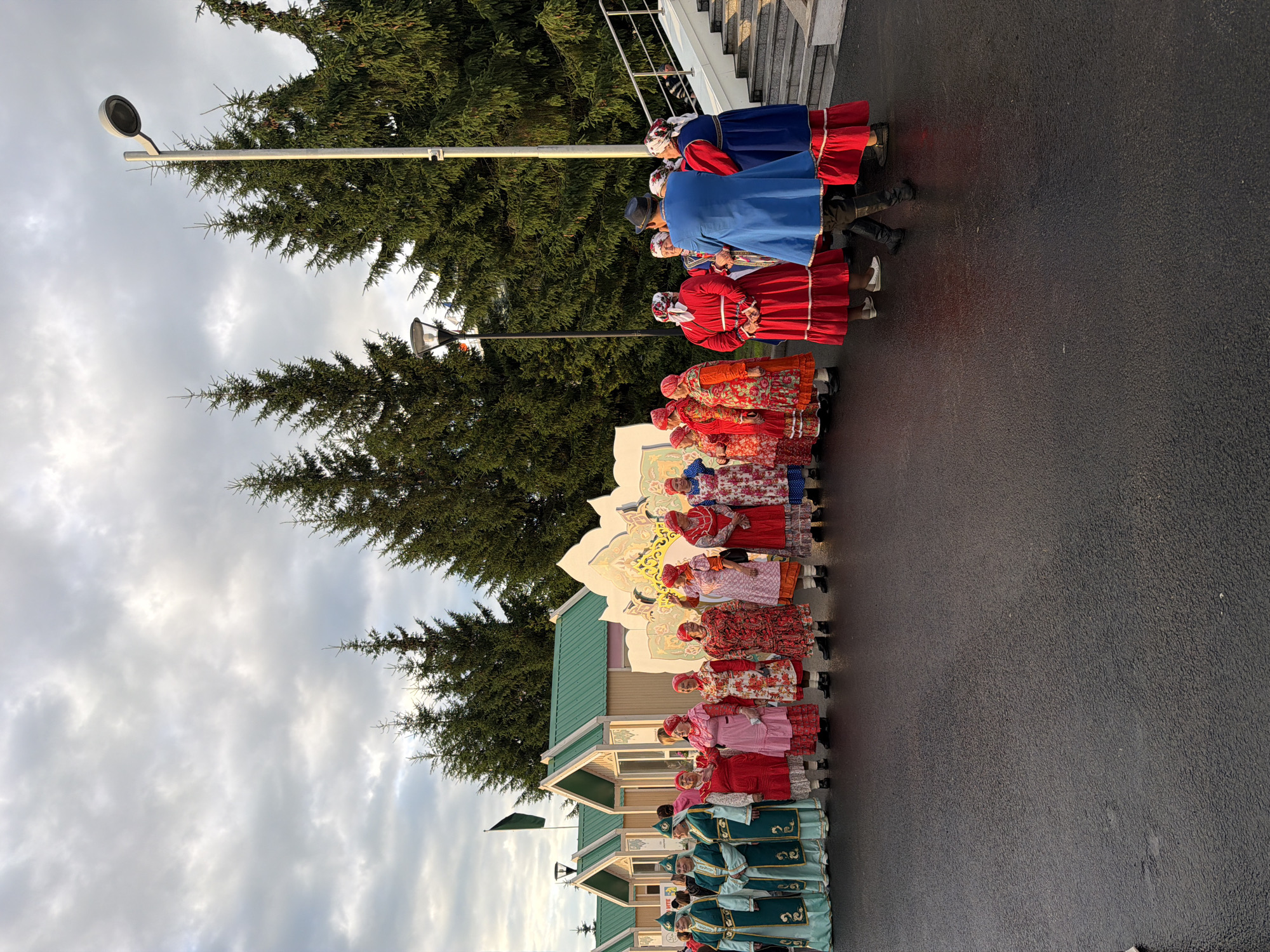 A group of people in traditional clothing stand in front of a row of small, colorful buildings, with trees and a cloudy sky in the background. The people are lined up and appear to be posing for a photograph. Some of the people are women wearing long, brightly colored dresses and headscarves, while others are men wearing darker, more formal attire. The buildings have green roofs and light-colored walls, and they are arranged in a straight line. The trees are tall and evergreen, and they form a dense backdrop to the scene. The sky is overcast with clouds. The ground in front of the buildings is paved and appears to be wet, possibly from recent rain. There is a white decorative structure behind some of the people on the right side of the image. A tall lamp post is visible on the right side of the image, and there are stairs leading up to a higher level behind the group on the right.