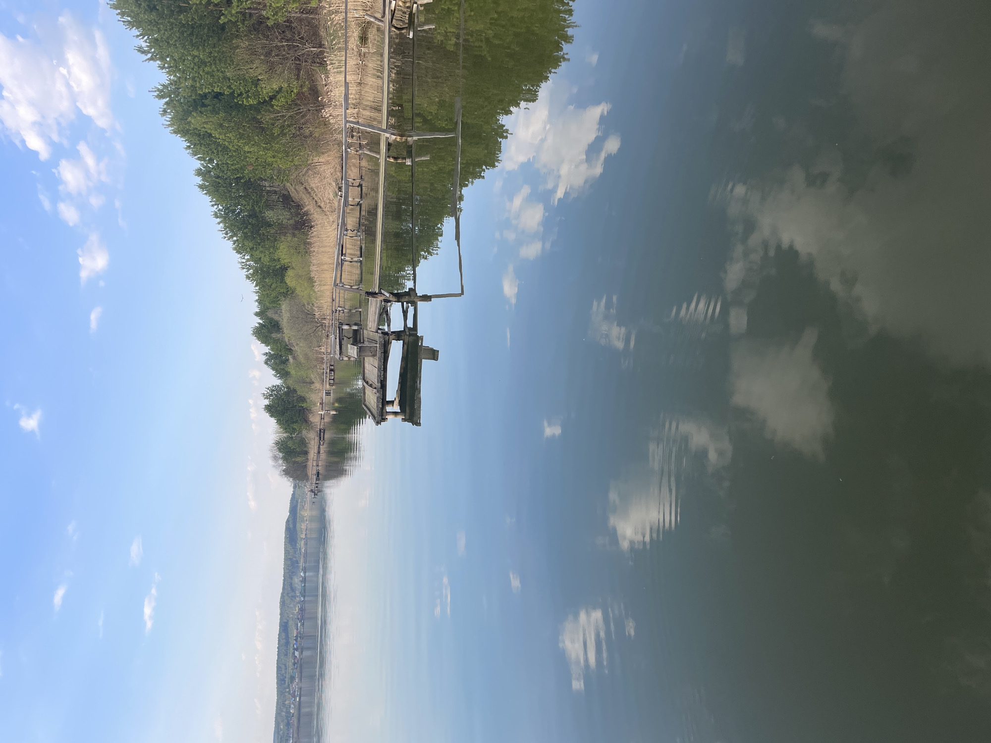 A landscape photograph taken in Suksun, Russia, shows a horizontal orientation. A wooden dock extends from the left side of the image into a body of water. A line of trees runs along the left edge of the image. The right side is dominated by the water, reflecting the sky and trees on the left. The sky is a clear blue with scattered white clouds.
