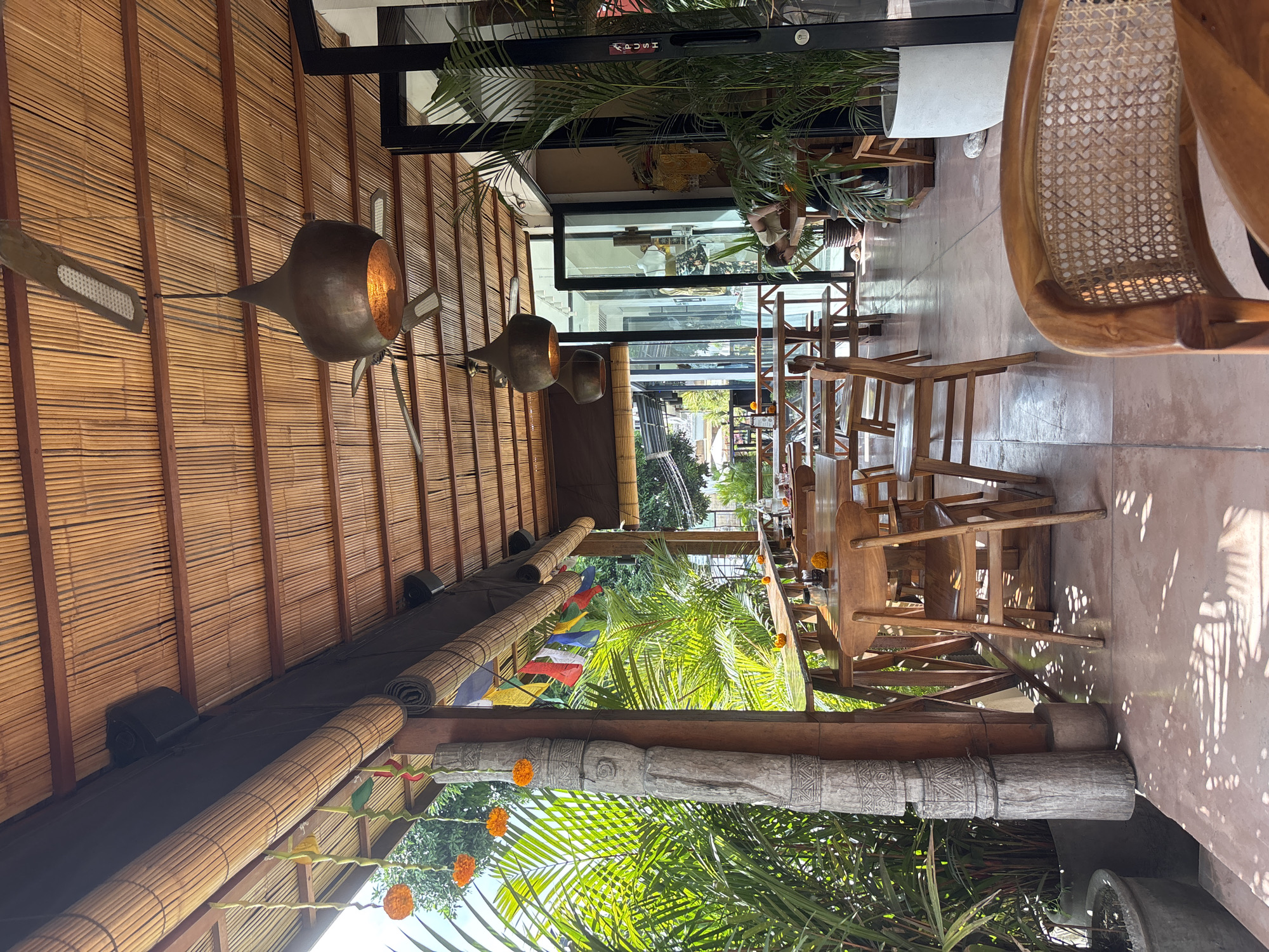 This appears to be the interior of a restaurant or covered patio in Kuta Selatan, Indonesia. The ceiling and walls are constructed with bamboo. Hanging from the ceiling are three metal light fixtures. Tables and wooden chairs are arranged along the length of the space. Beyond the open side of the structure are trees and foliage. On the outside, there is a decorated wooden beam. The restaurant is open to the outside, with green palms, trees, and colored flags. A figure can be seen through a glass door in the back of the space.