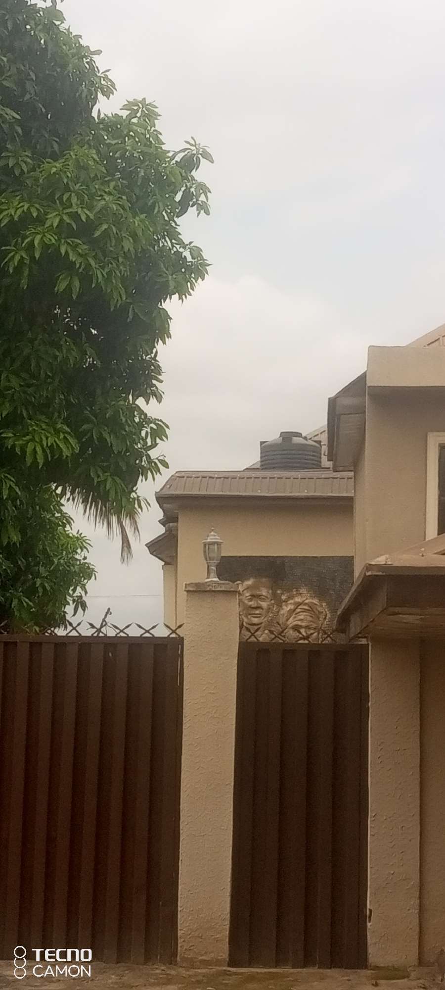 The image captures a section of a beige-colored building's exterior in Jalingo, Nigeria. A dark brown corrugated metal fence runs across the foreground, topped with barbed wire. To the left, a large tree with dense green foliage dominates the frame. The building itself has a light brown roof and a water tank is visible on its upper level. A decorative relief featuring two sculpted faces is mounted on a beige pillar near the fence. A lantern-style light fixture is positioned above the relief. The sky is overcast. The Tecno Camon logo is present in the bottom left corner.