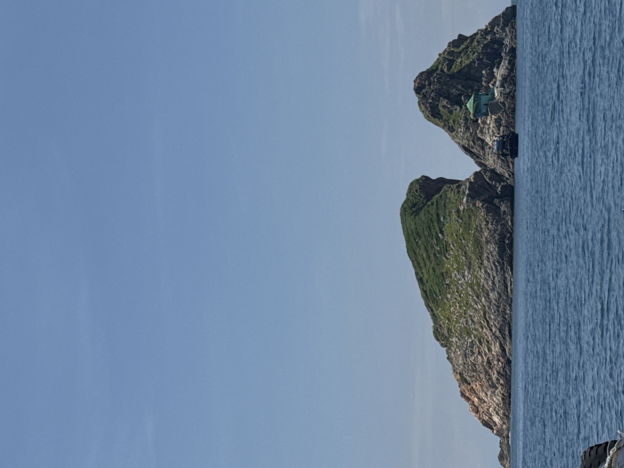 The image shows two rocky islands in the ocean, partially covered in green vegetation. A small, light green building with a dark roof is situated on the side of the right island, near the water. A dark-colored boat is anchored in front of the right island. The sky is clear and blue. The scene appears to be of a natural seascape.